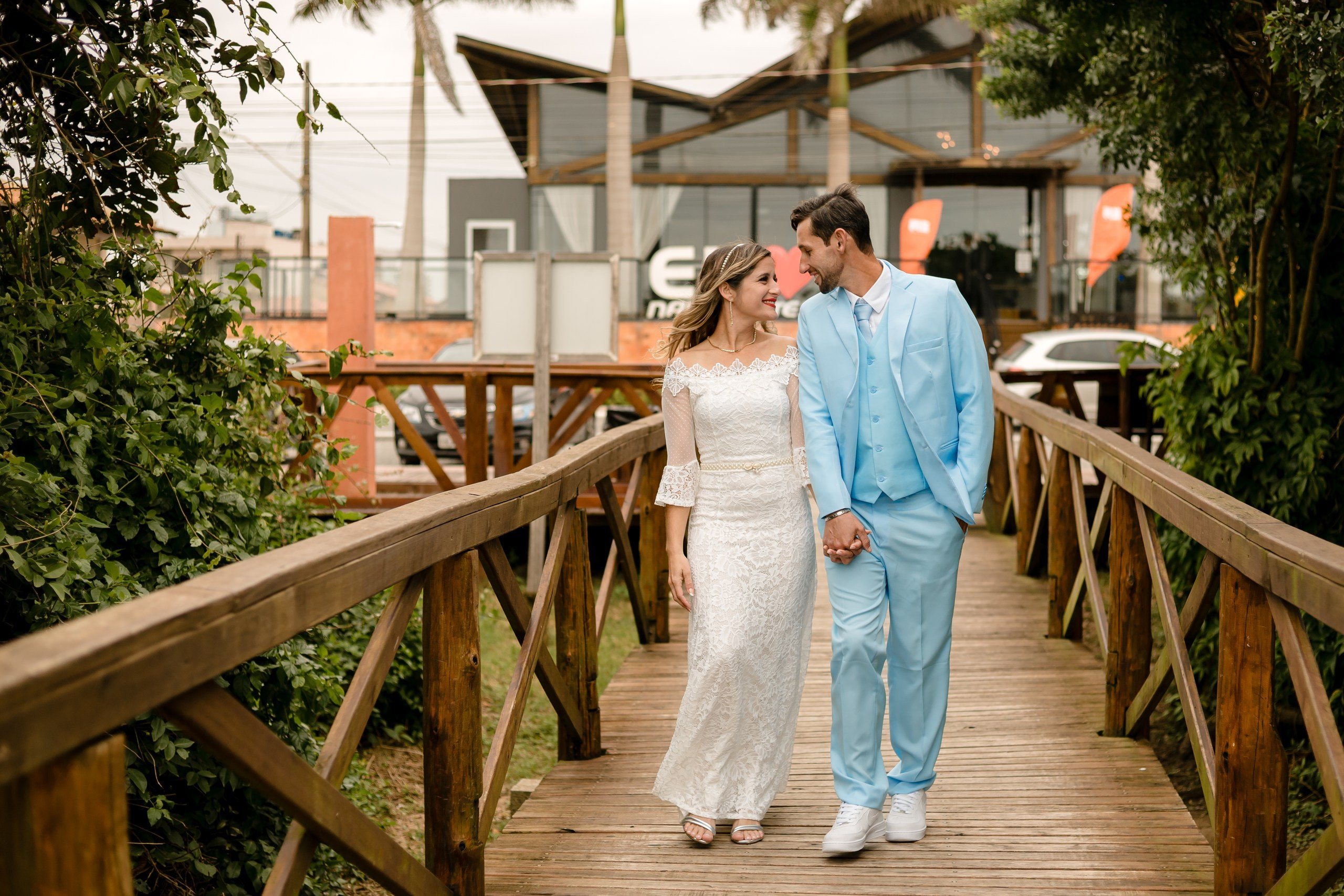 Fotógrafo de casamento em Navegantes; o casal de mãos dadas, caminhando e trocando olhares apaixonados sobre uma ponte de madeira cercada por vegetação durante ensaio externo.