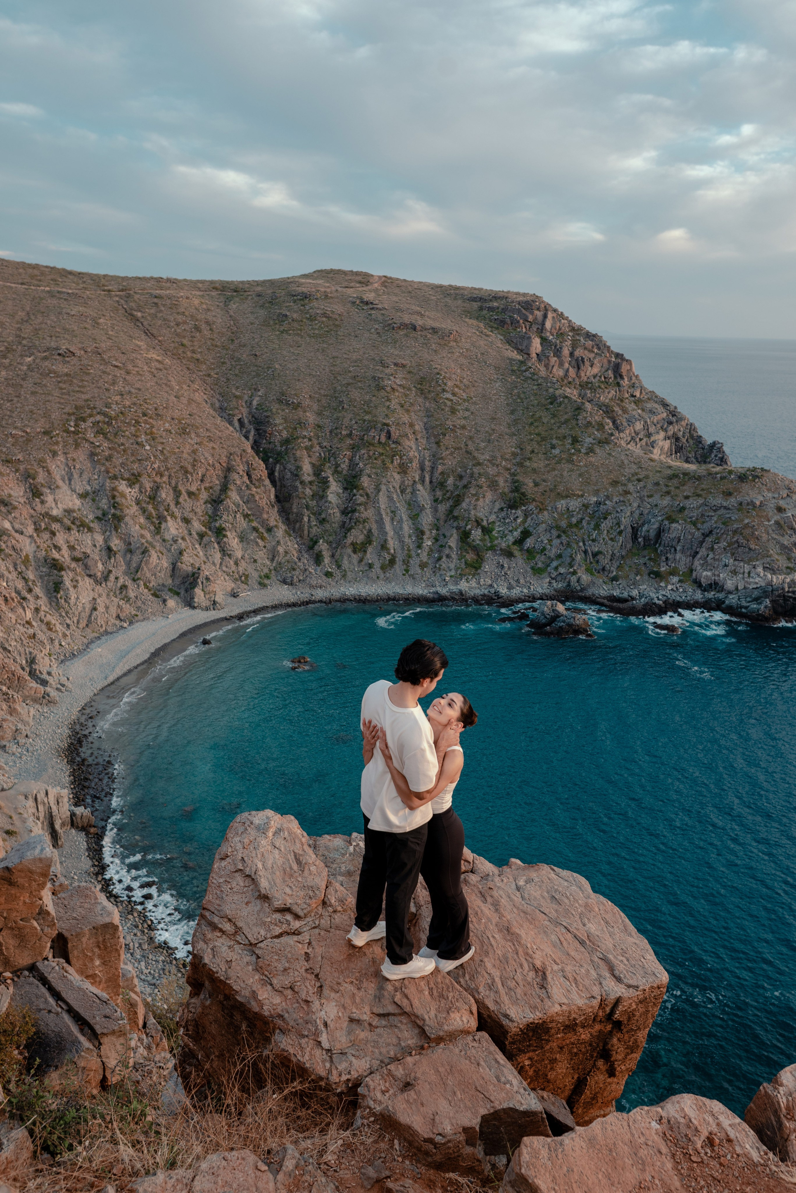 Engaged couple embracing on cliff edge overlooking stunning turquoise cove at Punta Lobos, Todos Santos Baja California Sur