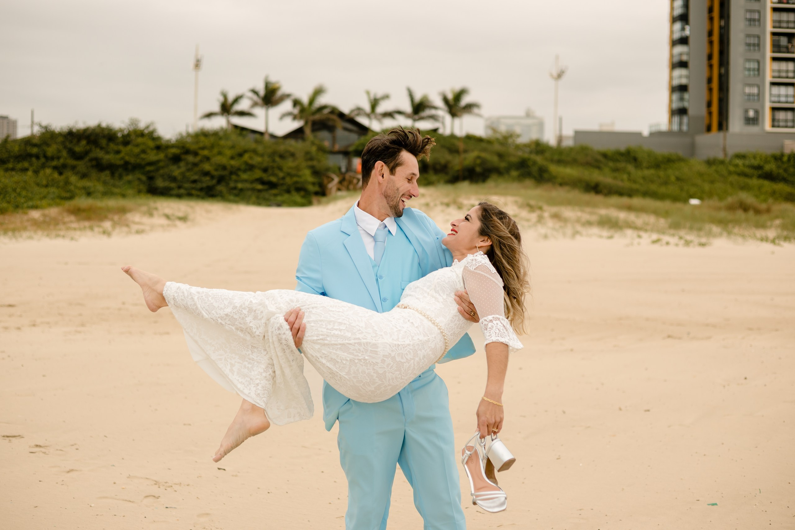 Fotógrafo de casamento em Navegantes; o noivo pega a noiva ao colo na areia da praia, ambos sorrindo enquanto ela segura os sapatos durante o ensaio externo.