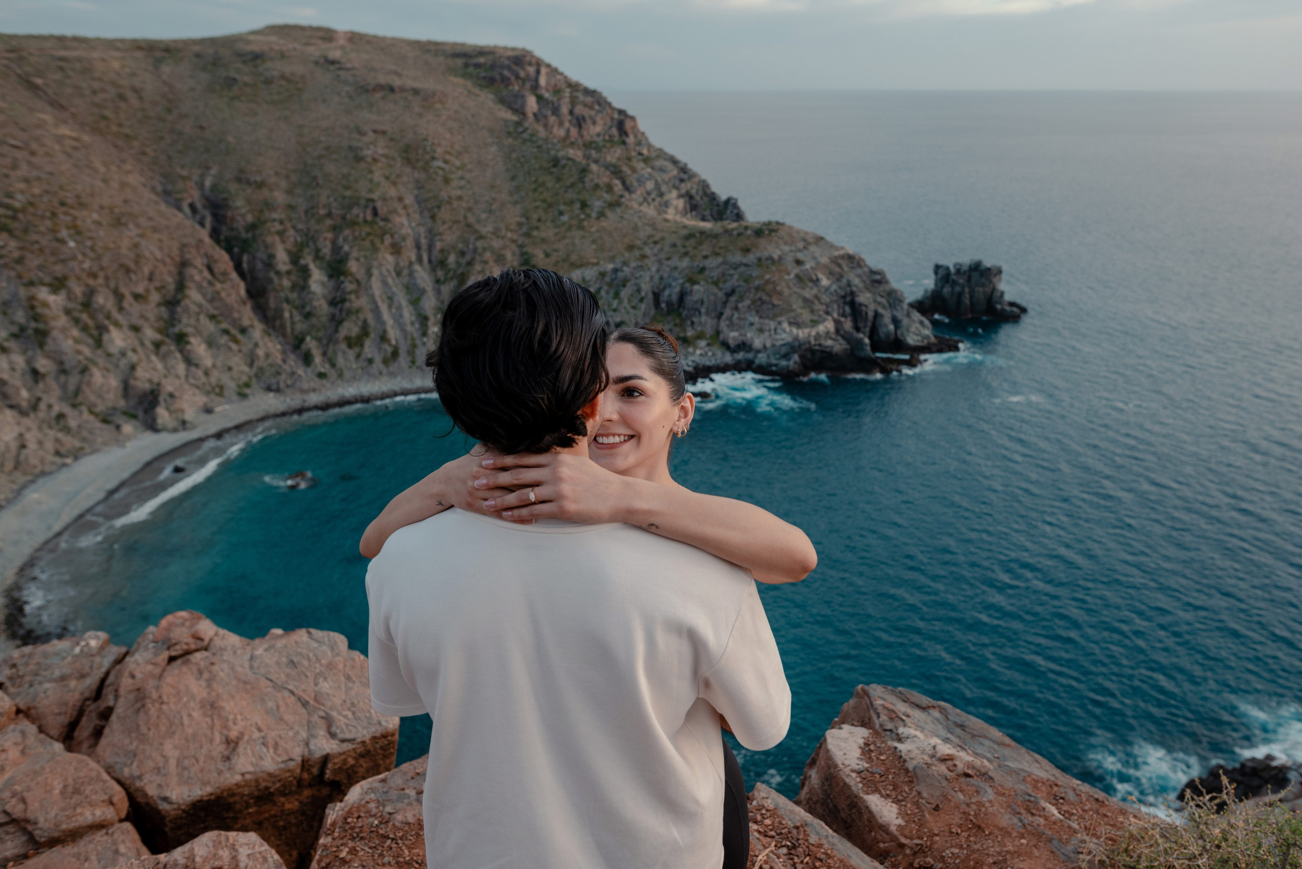 Woman hugging man from behind overlooking Punta Lobos bay and cliffs during engagement session, Todos Santos Baja California Sur