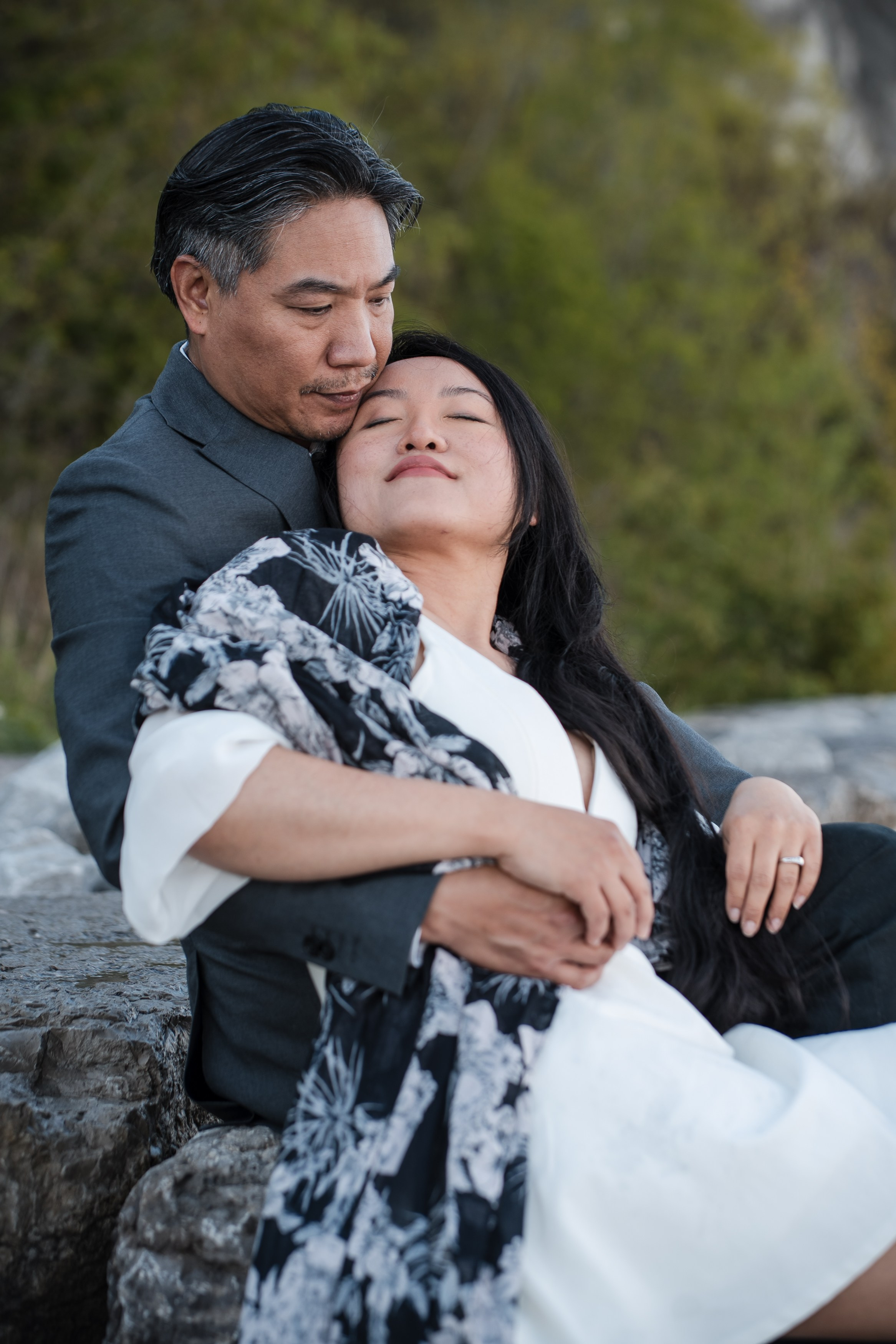 An engaged couple sits on a rocky shore.