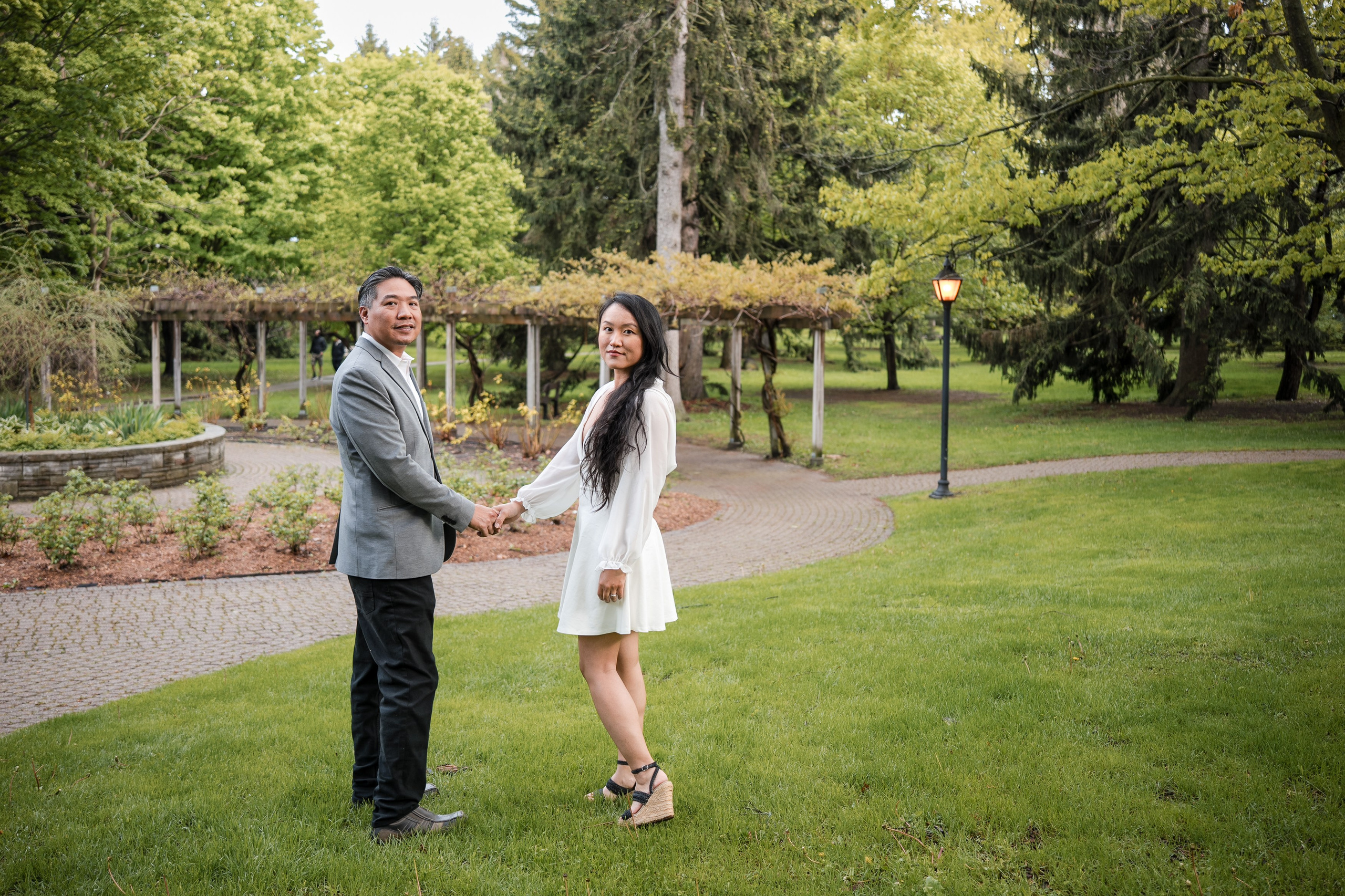 Engaged couple holding hands during a photoshoot in a park surrounded by green trees and a garden arbor.