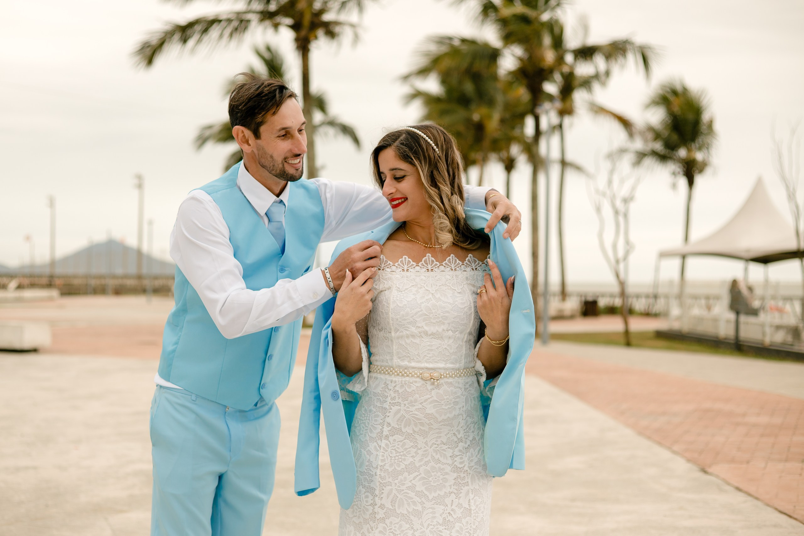 Fotógrafo de casamento em Navegantes; o noivo coloca o paletó sobre os ombros da noiva sorridente durante ensaio romântico na orla da praia.