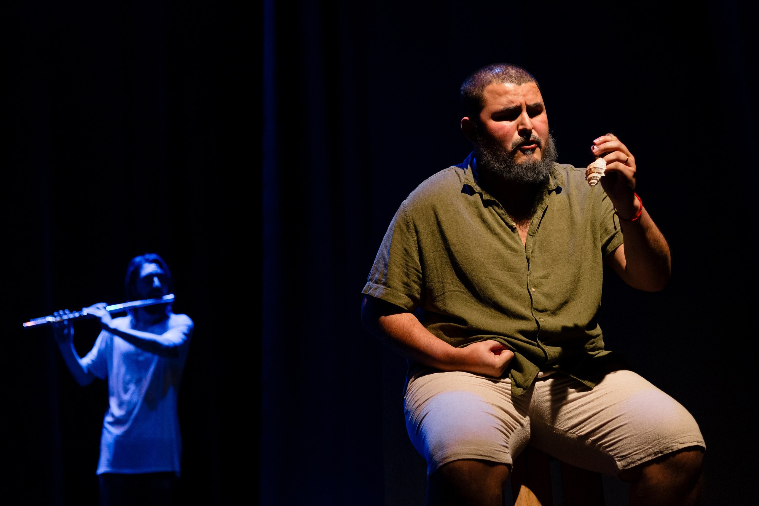 Apresentação teatral com homem segurando objeto enquanto outro aparece atrás tocando instrumento musical.