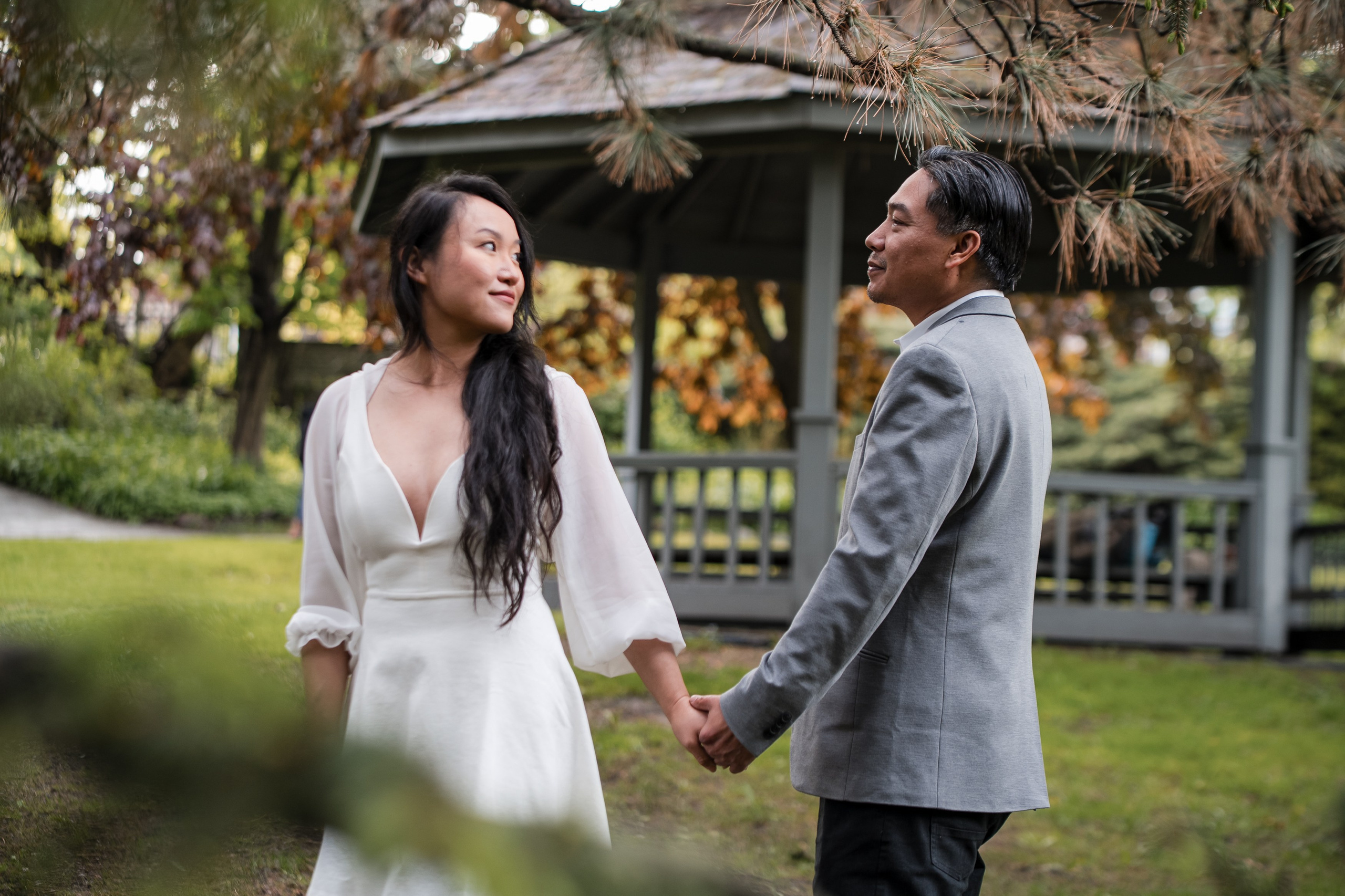 Engaged couple holding hands during a photoshoot in a park surrounded by green trees and a garden arbor.