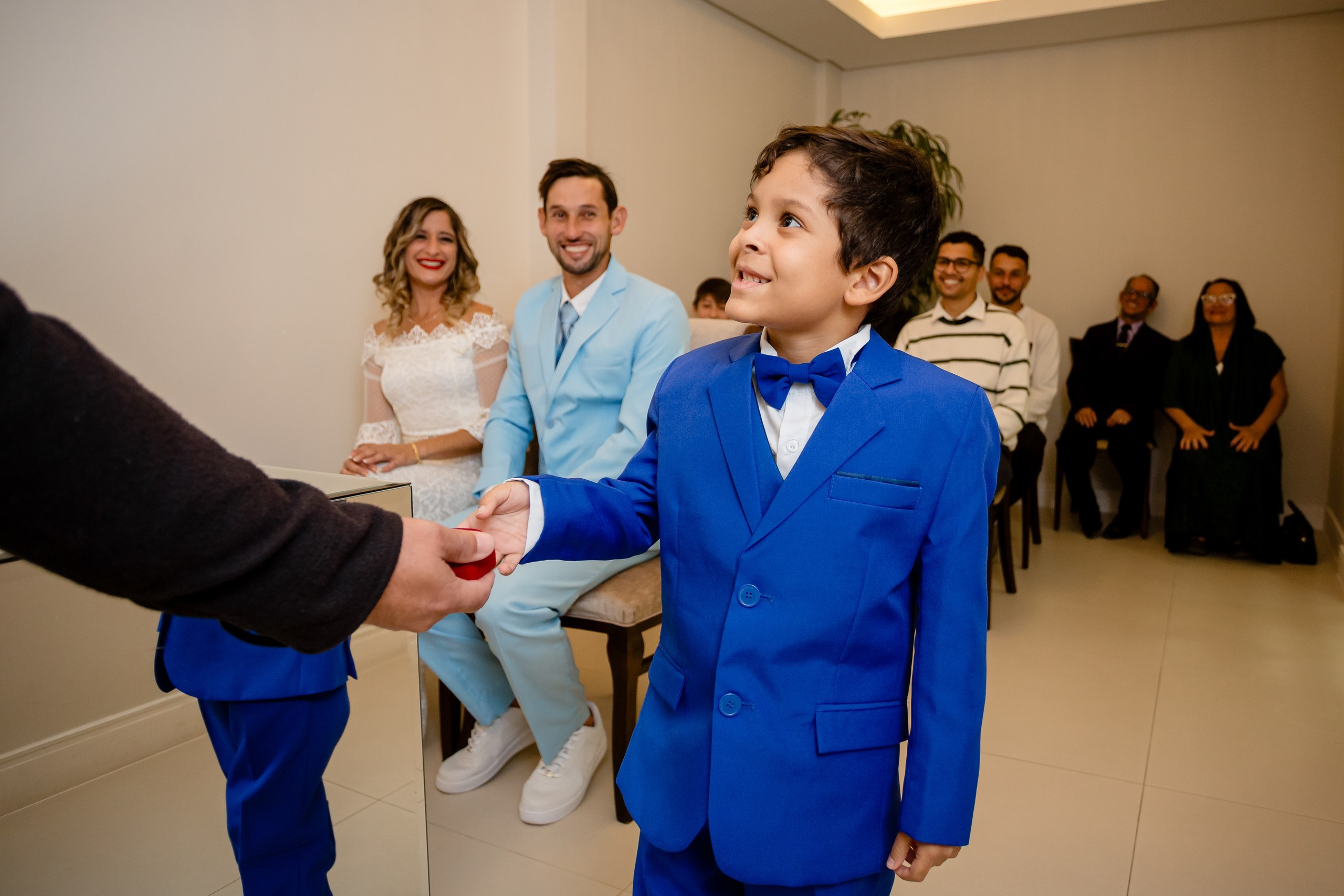 Fotógrafo de casamento civil em Navegantes; pajem de terno azul sorrindo ao receber as alianças durante a cerimônia, com os noivos ao fundo.