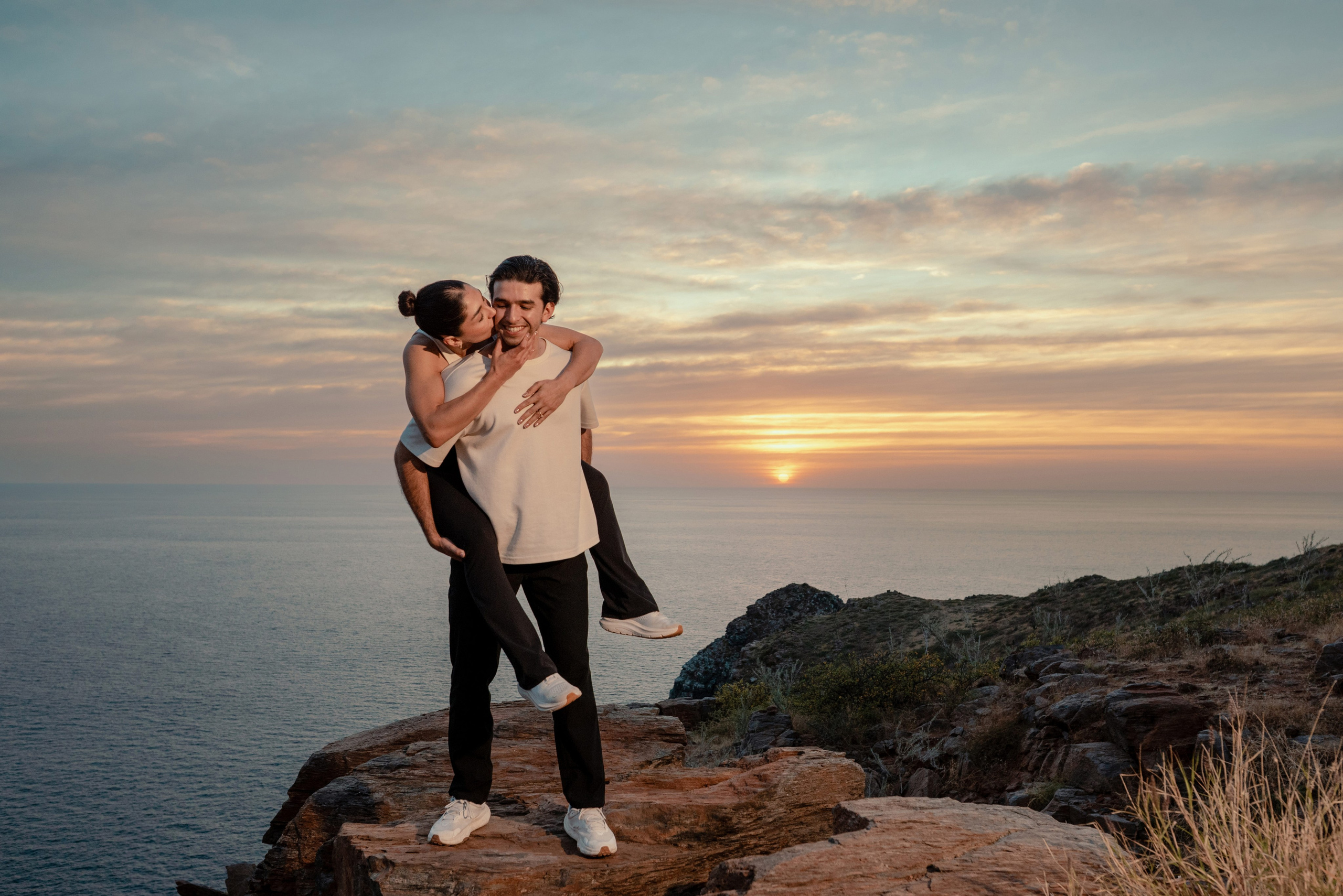 Couple laughing as man carries woman on his back at Punta Lobos cliffs during golden hour sunset, Todos Santos Baja California Sur