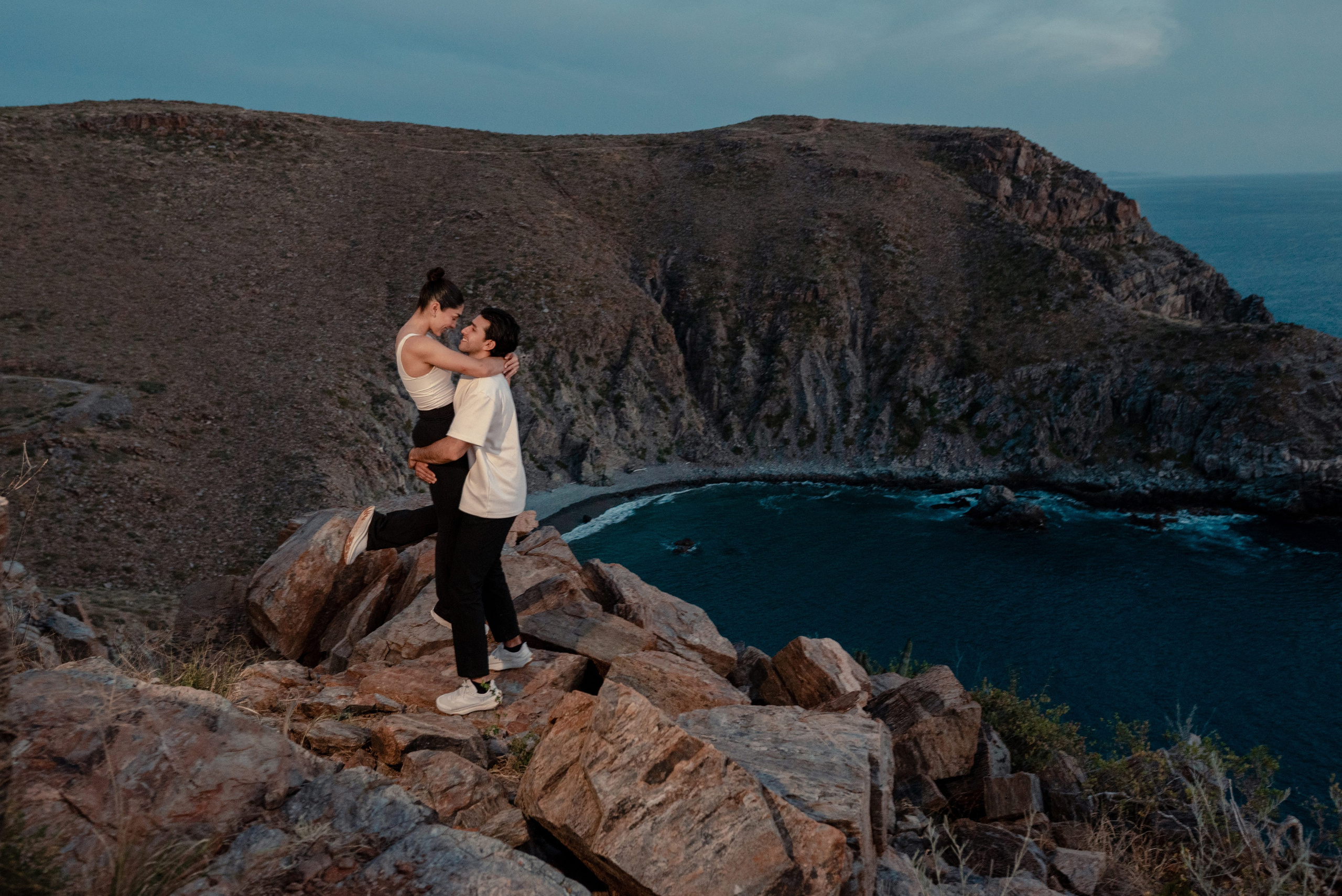 Couple kissing on rocky cliffs at Punta Lobos with turquoise bay and dramatic coastline at dusk, Todos Santos Baja California Sur