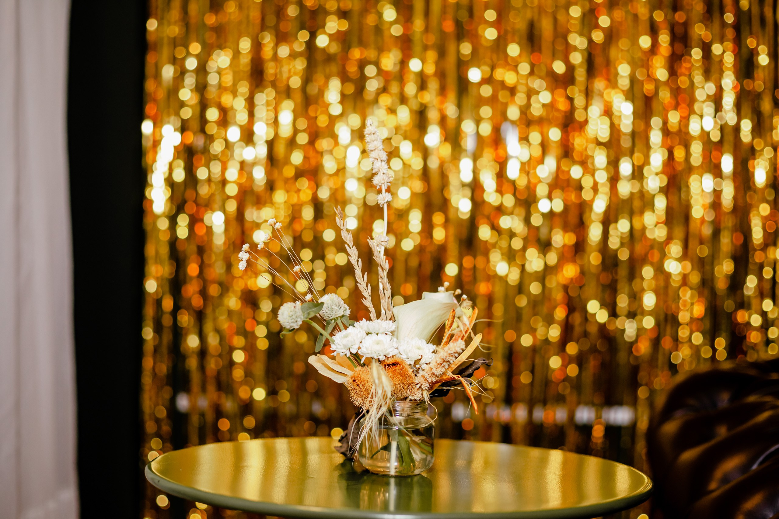 Fotógrafo festa de aniversário em Itajaí; arranjo floral decorativo sobre mesa de apoio em evento festivo com fundo iluminado por cortina de luzes.