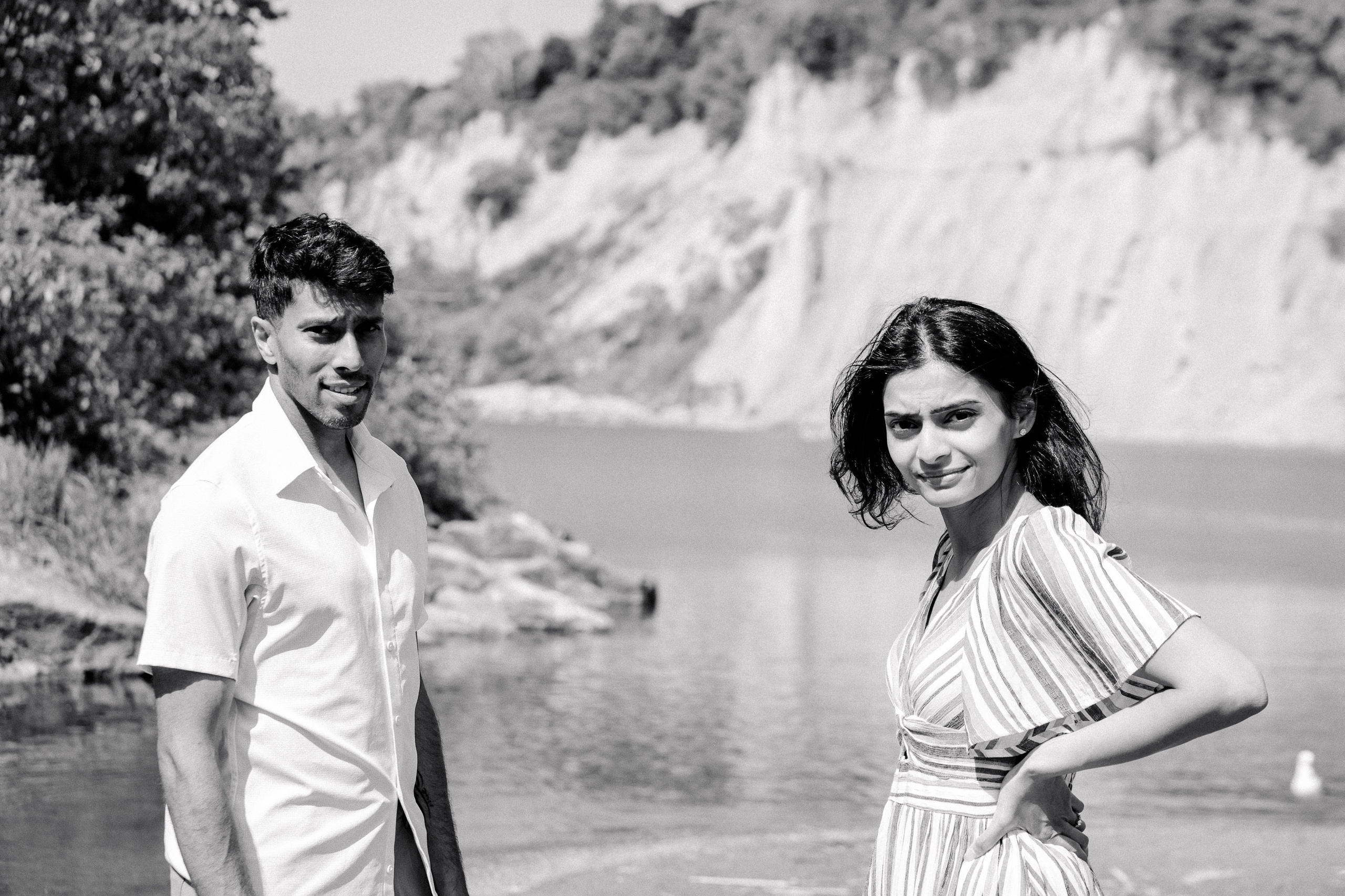 Black and white engagement photo of a couple by the water, with cliffs and trees in the background — capturing a candid and serene moment in nature.