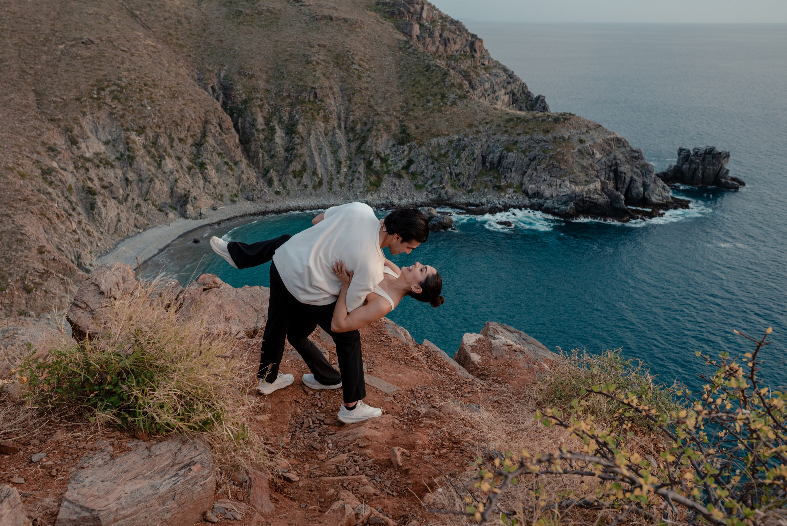 Groom carrying fiancée in arms against golden sunset sky at Punta Lobos cliffs, Todos Santos Baja California Sur
