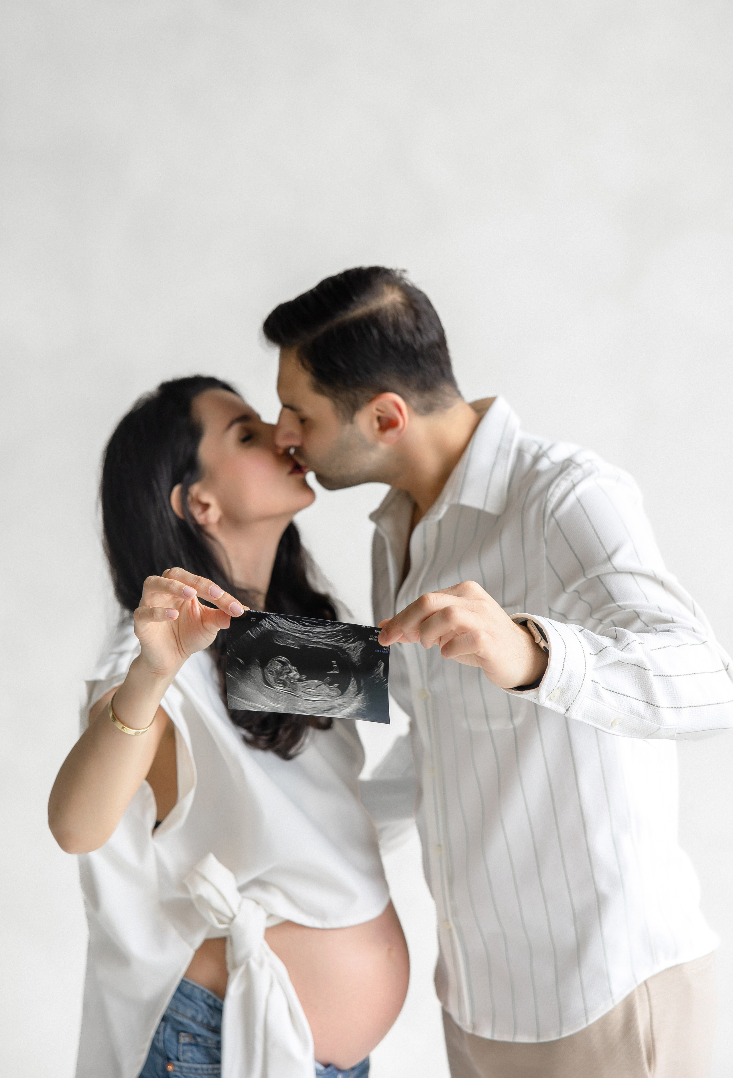 Pregnant couple kissing while holding ultrasound photo during maternity photoshoot in Toronto studio – elegant, minimalist pregnancy photography by Iana Banana