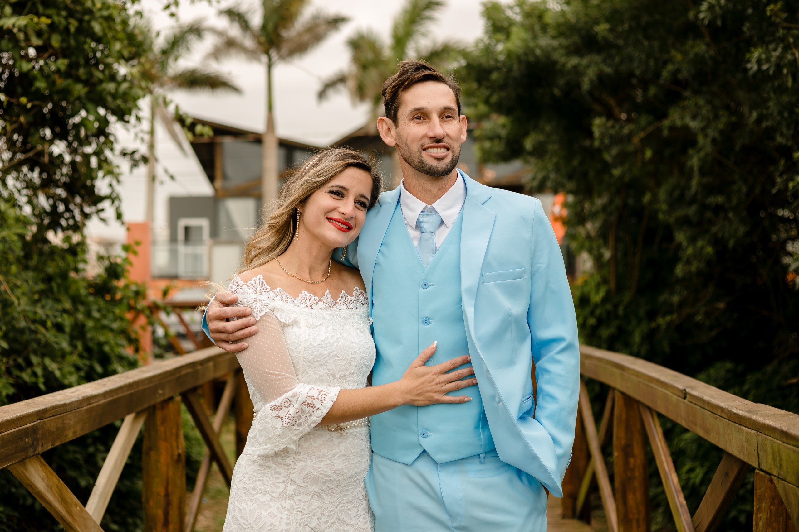 Fotógrafo de casamento em Navegantes; o casal abraçado na ponte de madeira cercada por vegetação verdejante durante ensaio externo na praia.
