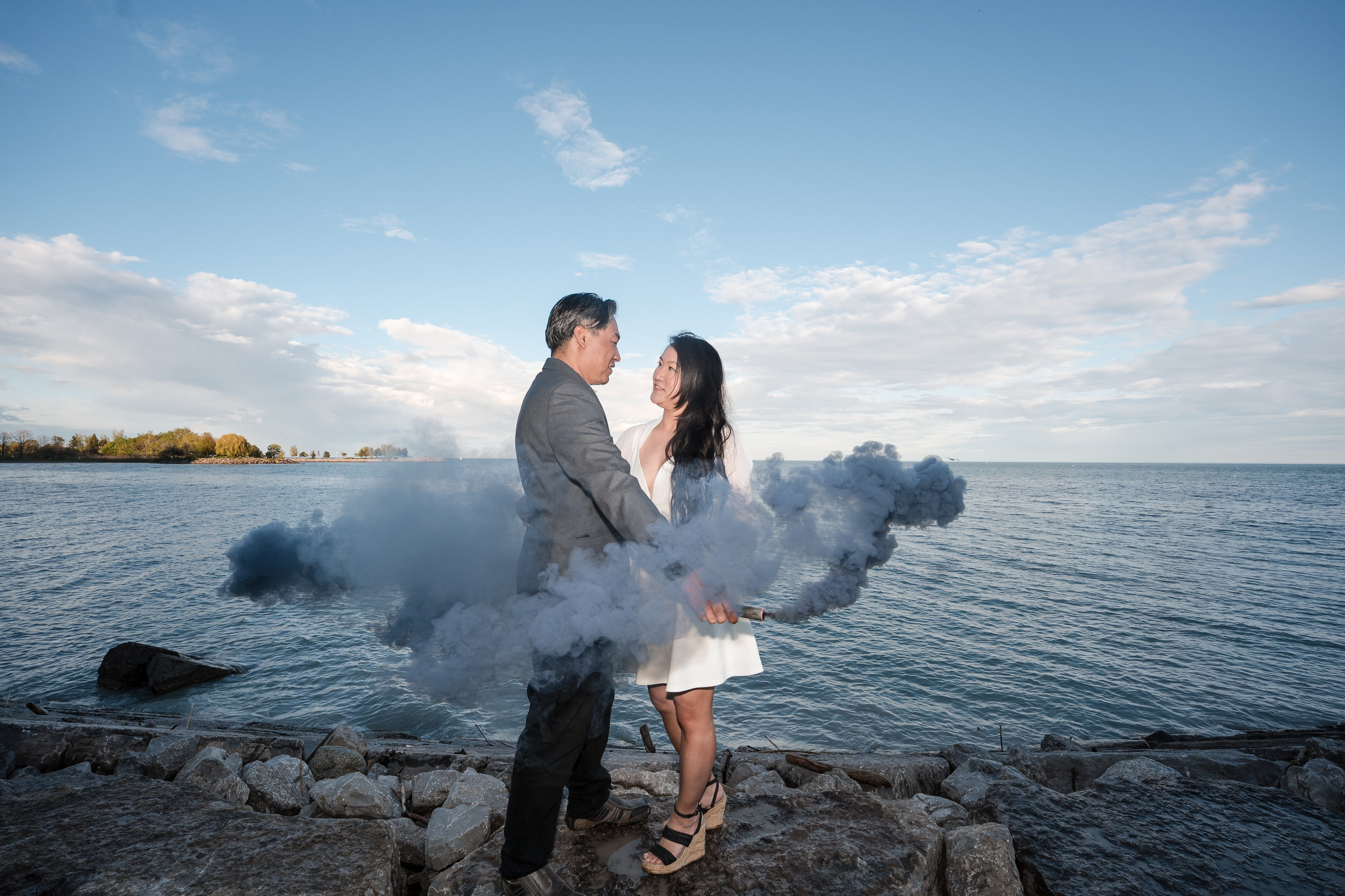 Engaged couple standing on rocky shore with smoke bomb, gazing at each other under a clear sky.