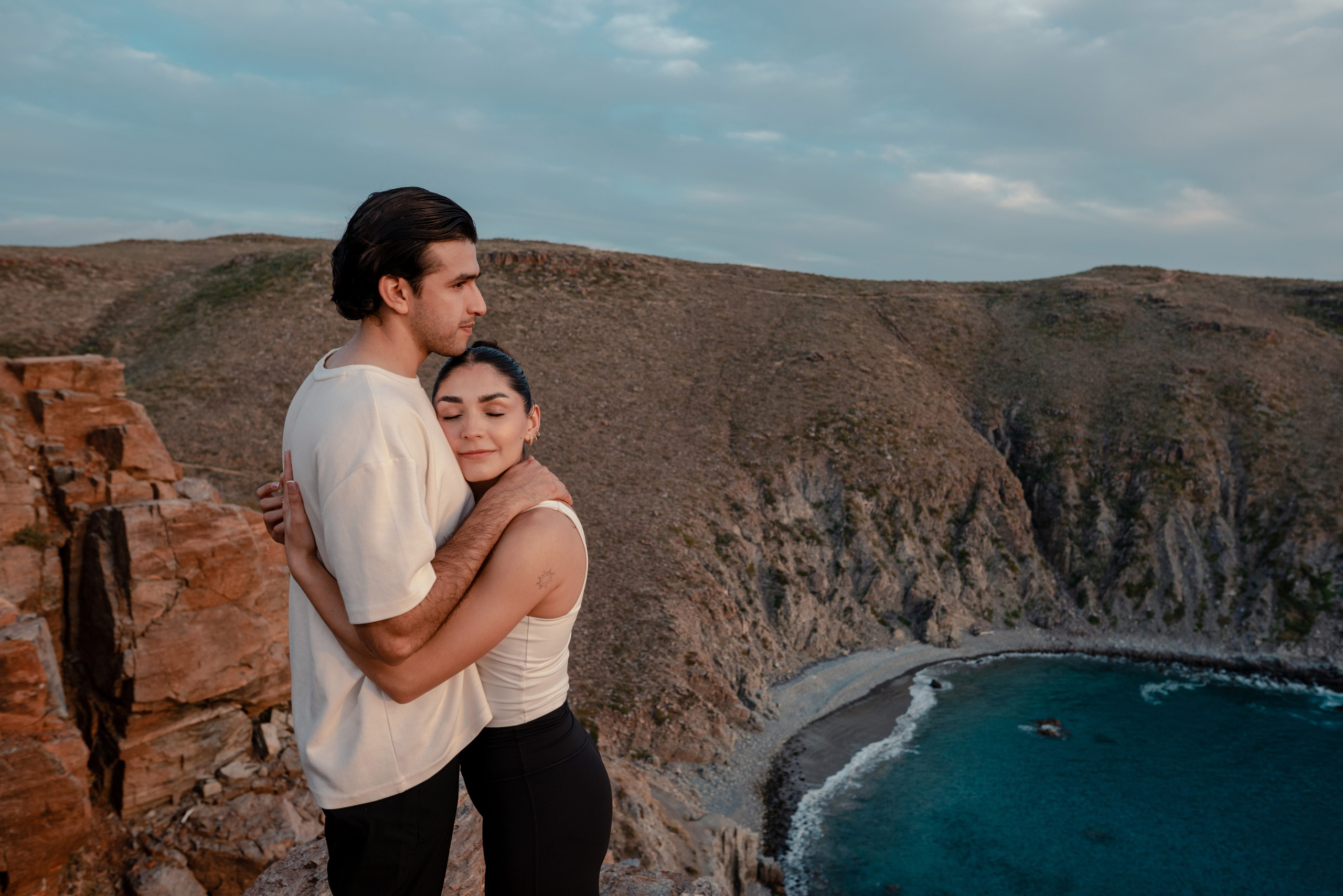 Couple embracing while looking over Punta Lobos cove with red earth cliffs and turquoise bay at sunset, Todos Santos Baja California Sur