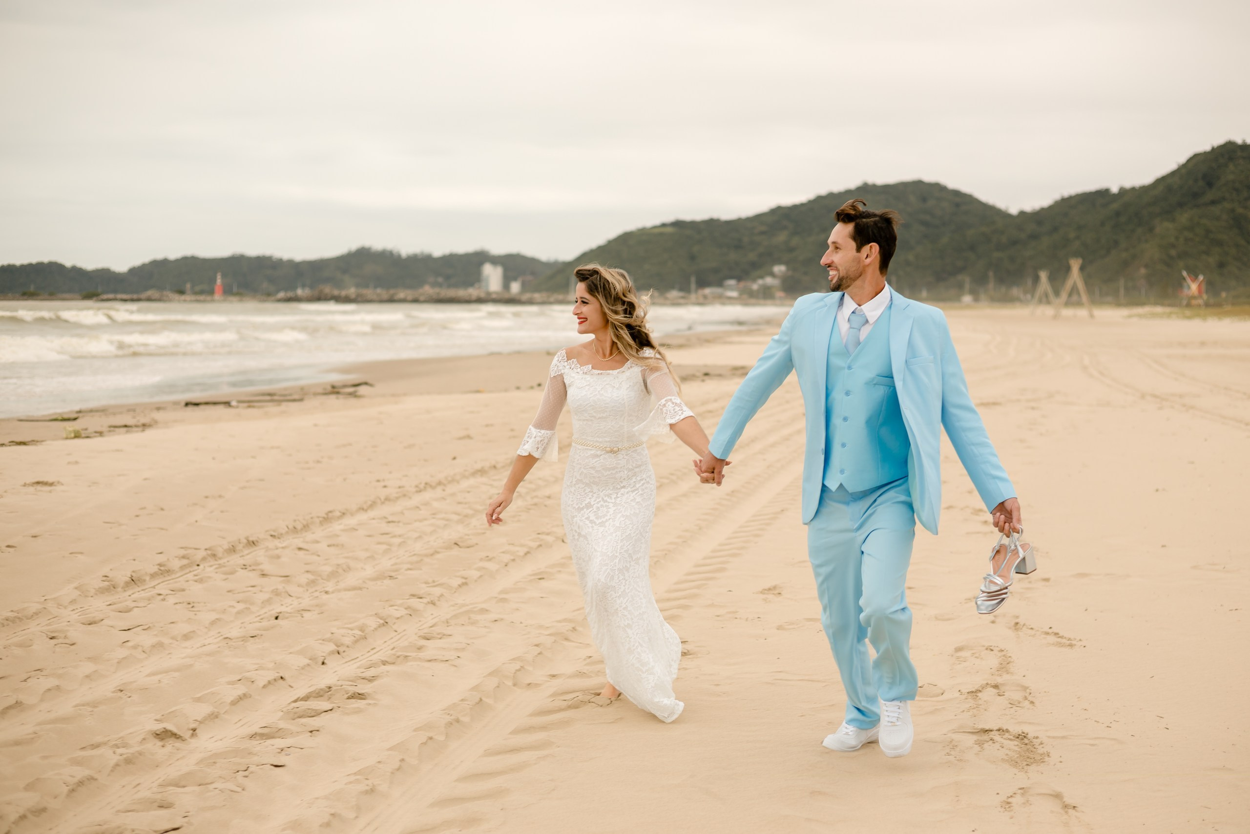 Fotógrafo de casamento em Navegantes; o casal corre de mãos dadas pela areia da praia, com a noiva descalça e o noivo segurando os sapatos, em um ensaio externo descontraído.