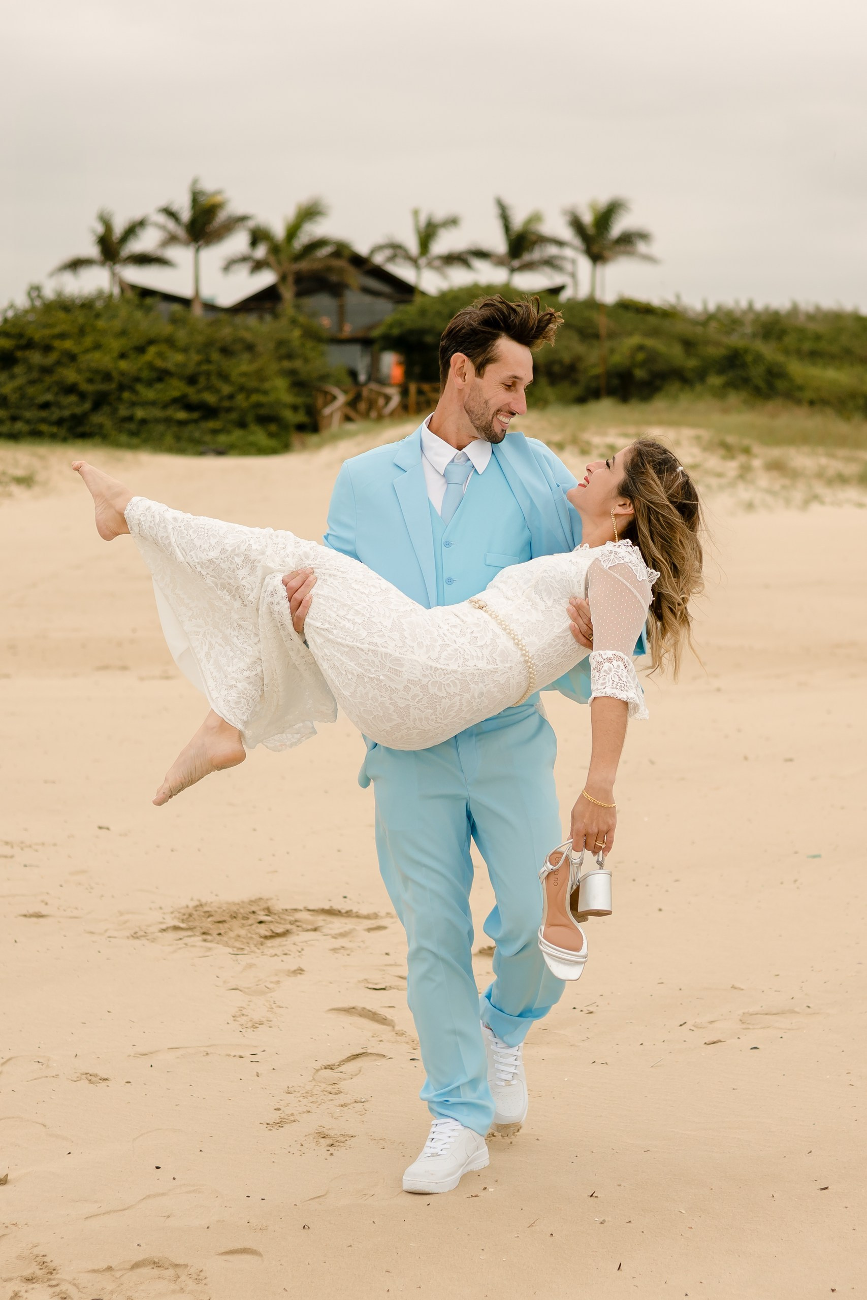 Fotógrafo de casamento em Navegantes; o noivo carrega a noiva ao colo enquanto caminham sorridentes pela areia da praia, tendo os sapatos na mão durante o ensaio externo.