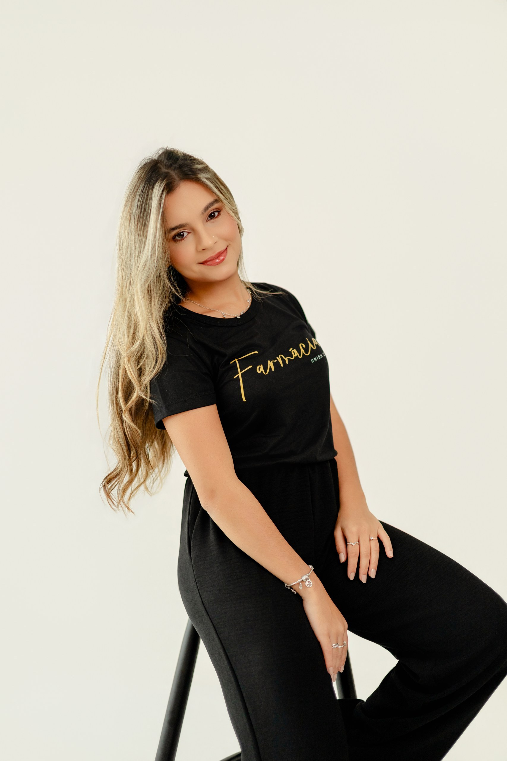 Fotografia de ensaio de estúdio em Balneário Camboriú com formanda de farmácia sentada, sorrindo e vestindo blusa preta personalizada.