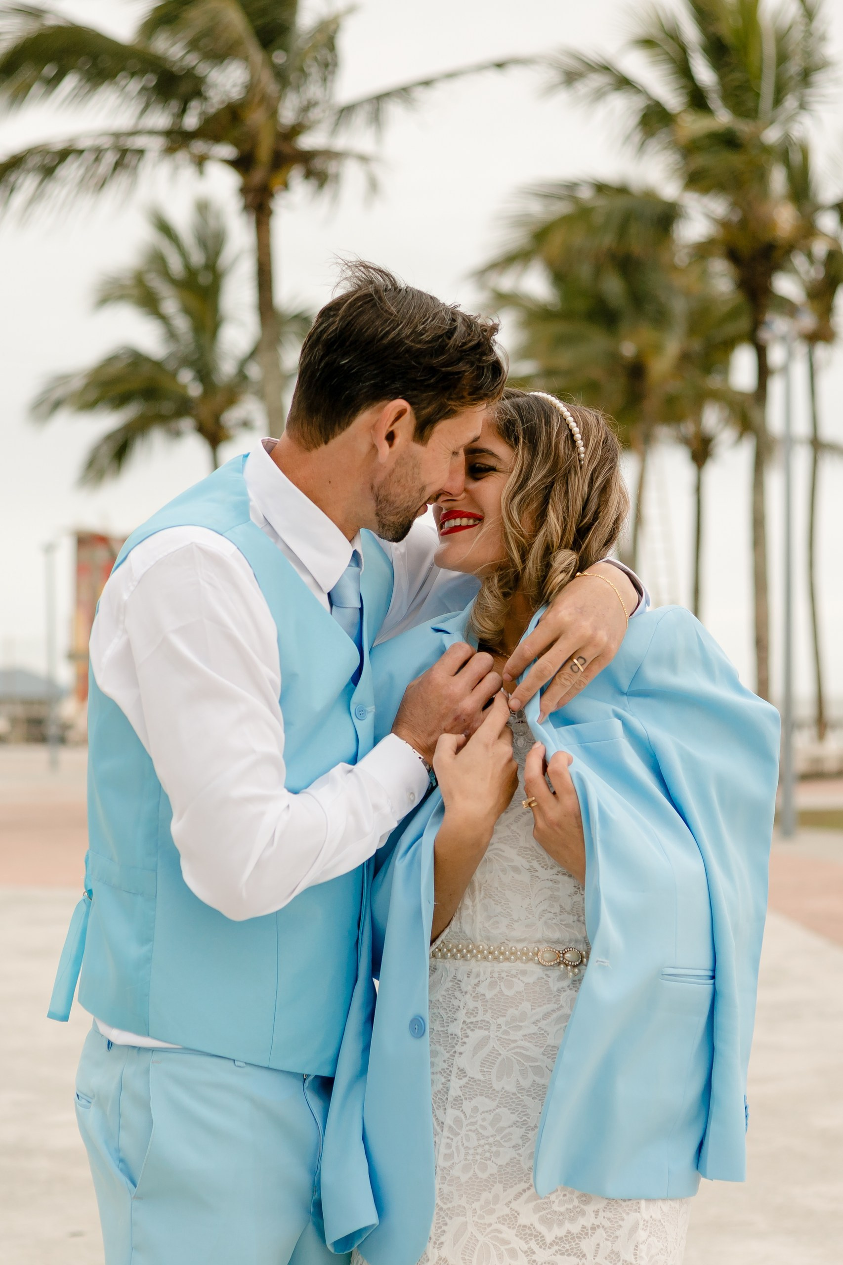 Fotógrafo de casamento em Navegantes; o noivo e a noiva trocam um sorriso apaixonado com palmeiras ao fundo durante ensaio de casal na praia.