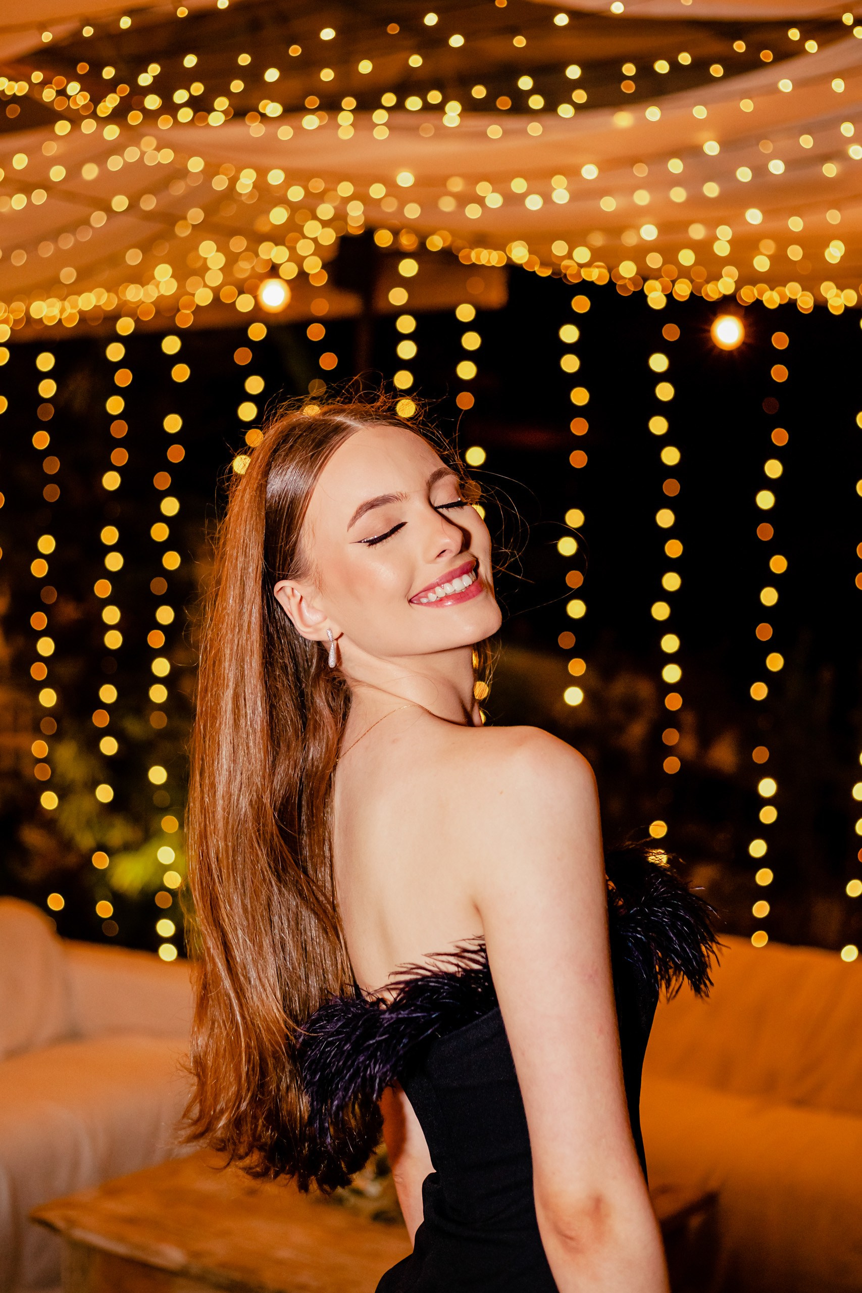 Fotógrafo festa de aniversário em Itajaí; retrato fechado e espontâneo da aniversariante sorrindo com os olhos fechados, tendo ao fundo a iluminação aconchegante de luzes suspensas.