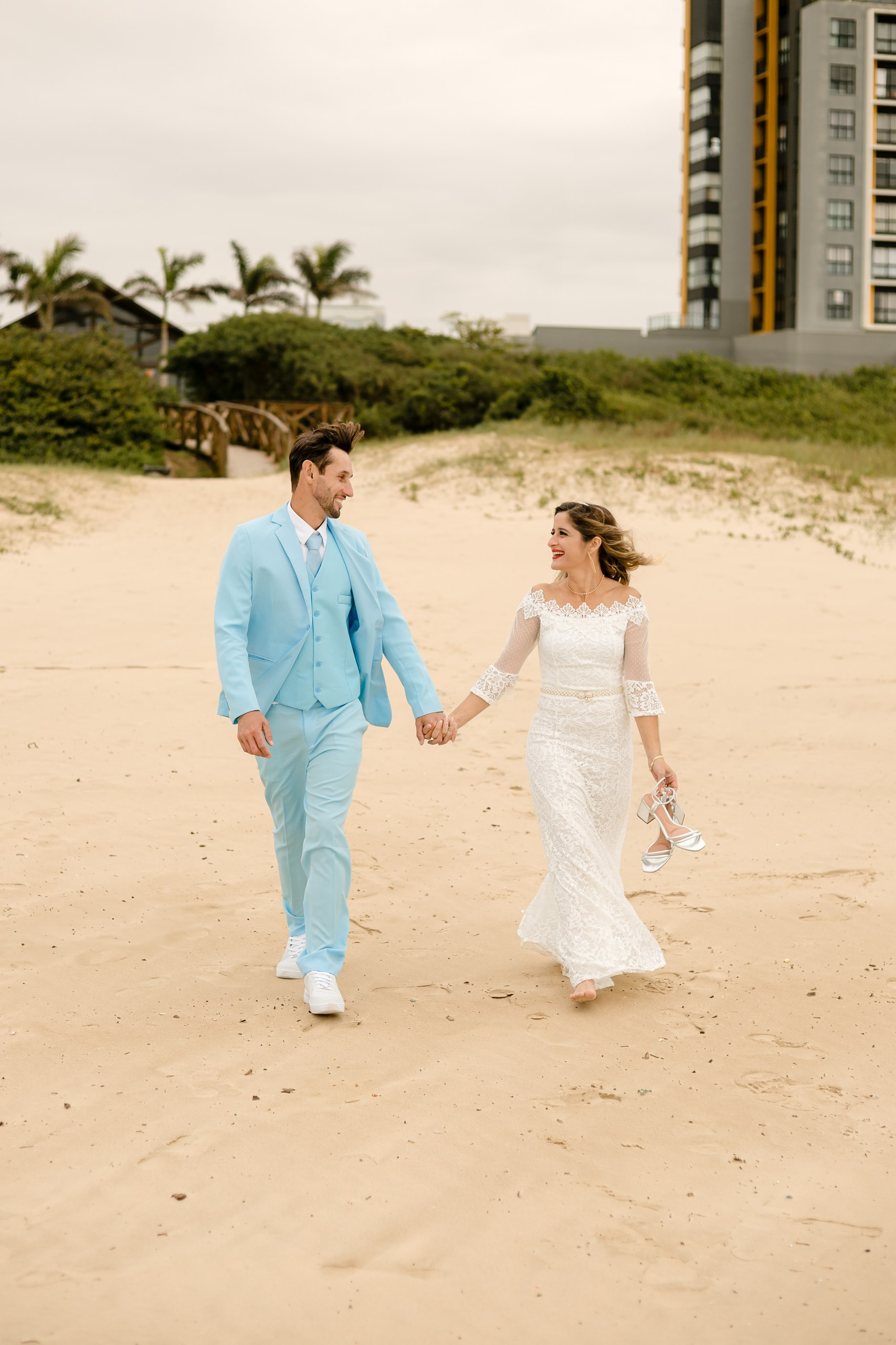 Fotógrafo de casamento em Navegantes; o casal caminha de mãos dadas descalço na areia da praia, com a noiva segurando os sapatos, durante ensaio fotográfico.