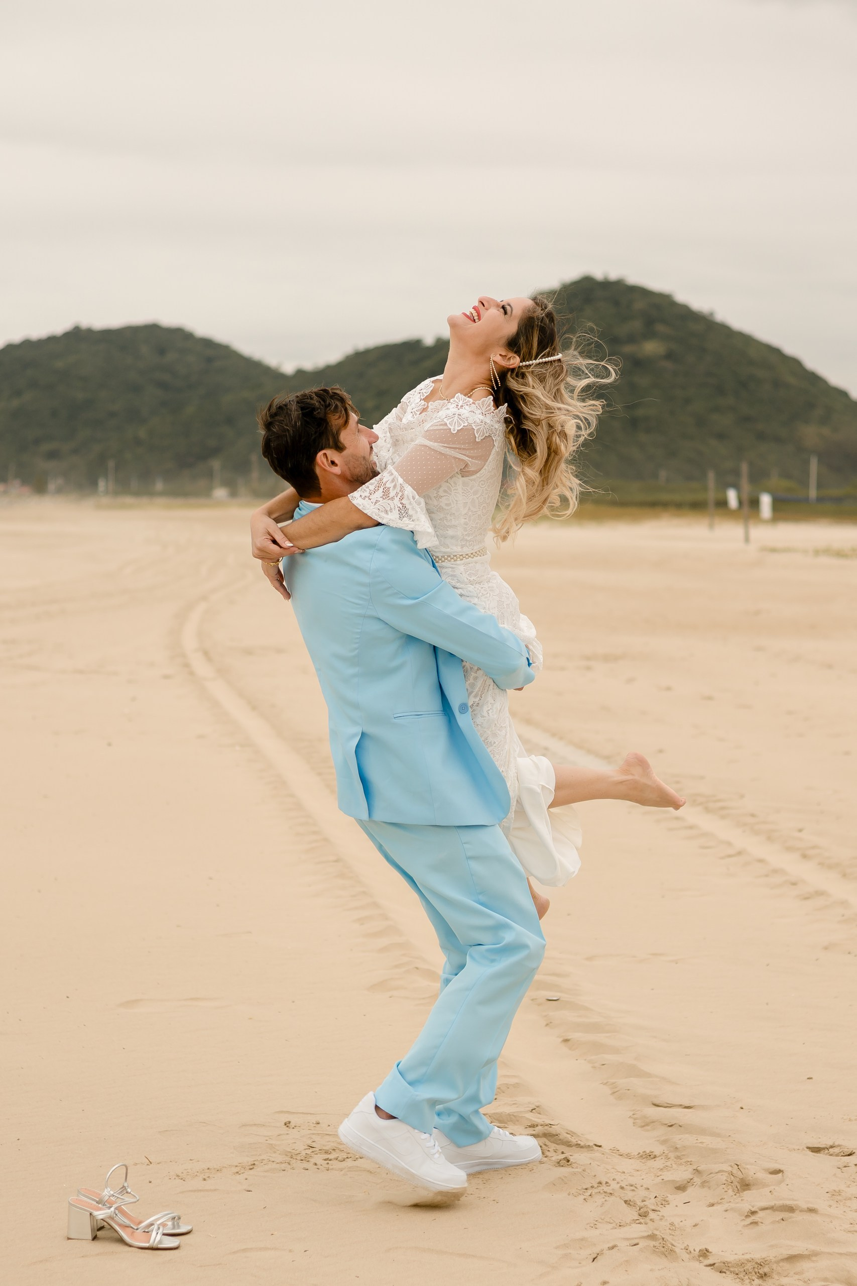 Fotógrafo de casamento em Navegantes; o noivo levanta a noiva no colo enquanto ela ri efusivamente, com os sapatos na areia ao lado, em ensaio externo na praia.