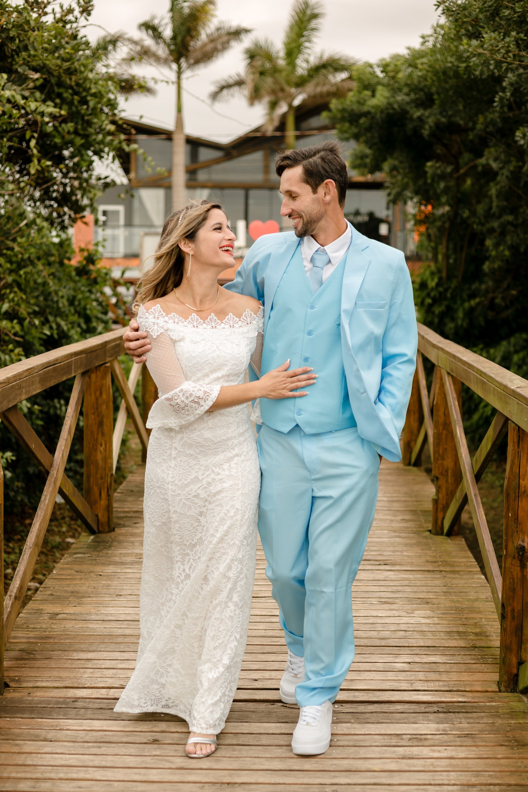 Fotógrafo de casamento em Navegantes; os recém-casados caminham sorridentes, de mãos dadas, sobre uma ponte de madeira cercada por vegetação e coqueiros durante ensaio externo.