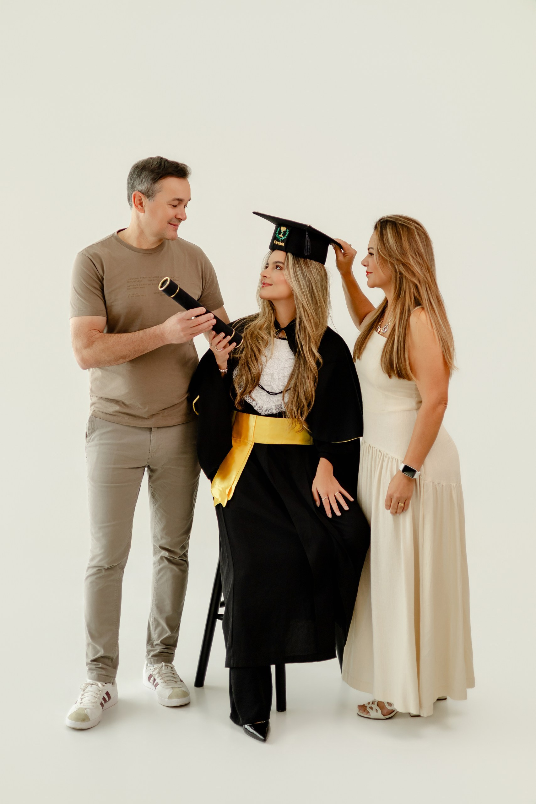 Fotografia de ensaio de formatura em Balneário Camboriú com formanda de farmácia sentada entre seus familiares, que seguram o canudo e ajustam o capelo.