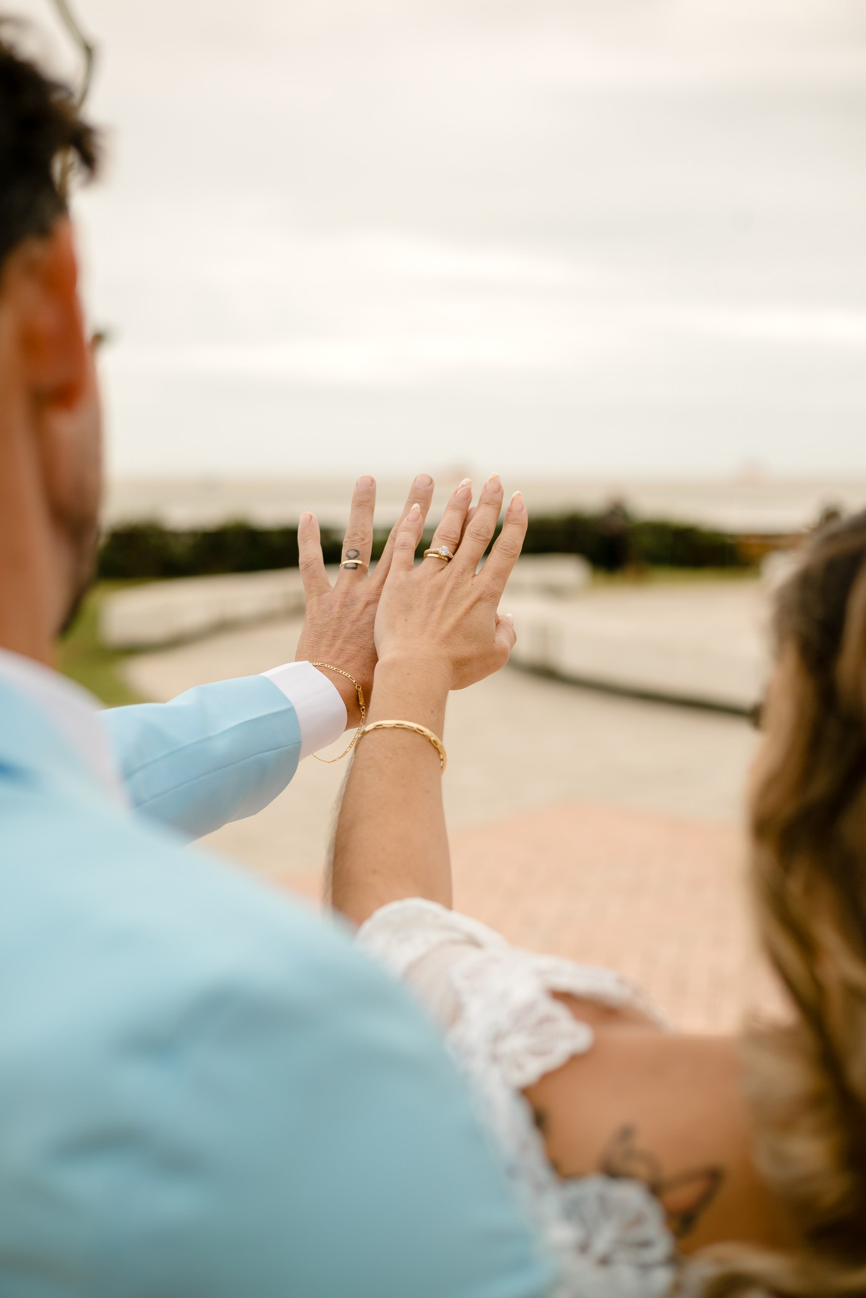 Fotógrafo de casamento em Navegantes; o casal mostrando as mãos com as alianças em primeiro plano e a paisagem da praia ao fundo durante ensaio fotográfico.