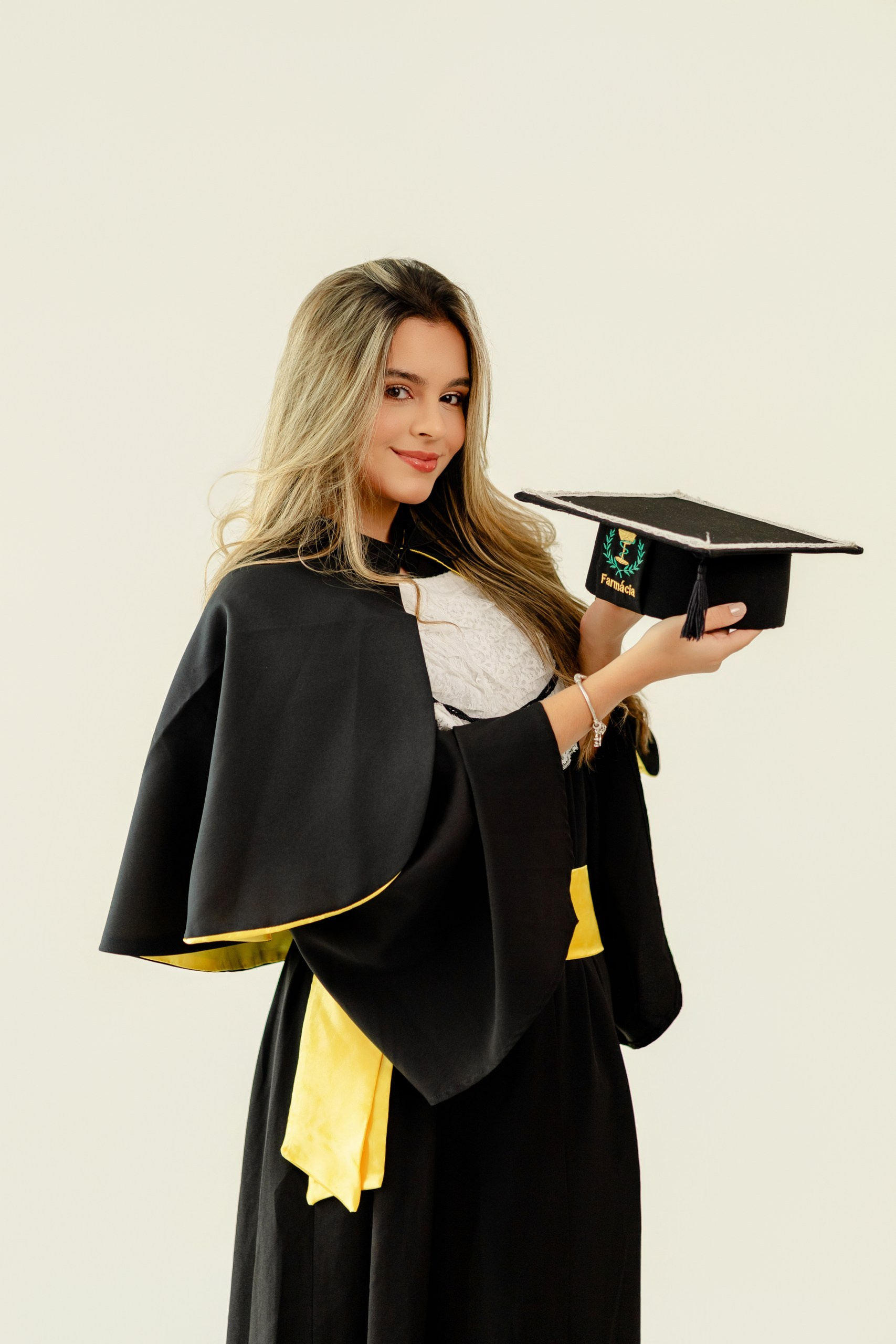 Fotografia de ensaio de formatura em Balneário Camboriú com formanda de farmácia vestindo beca preta com detalhes amarelos e segurando o capelo.