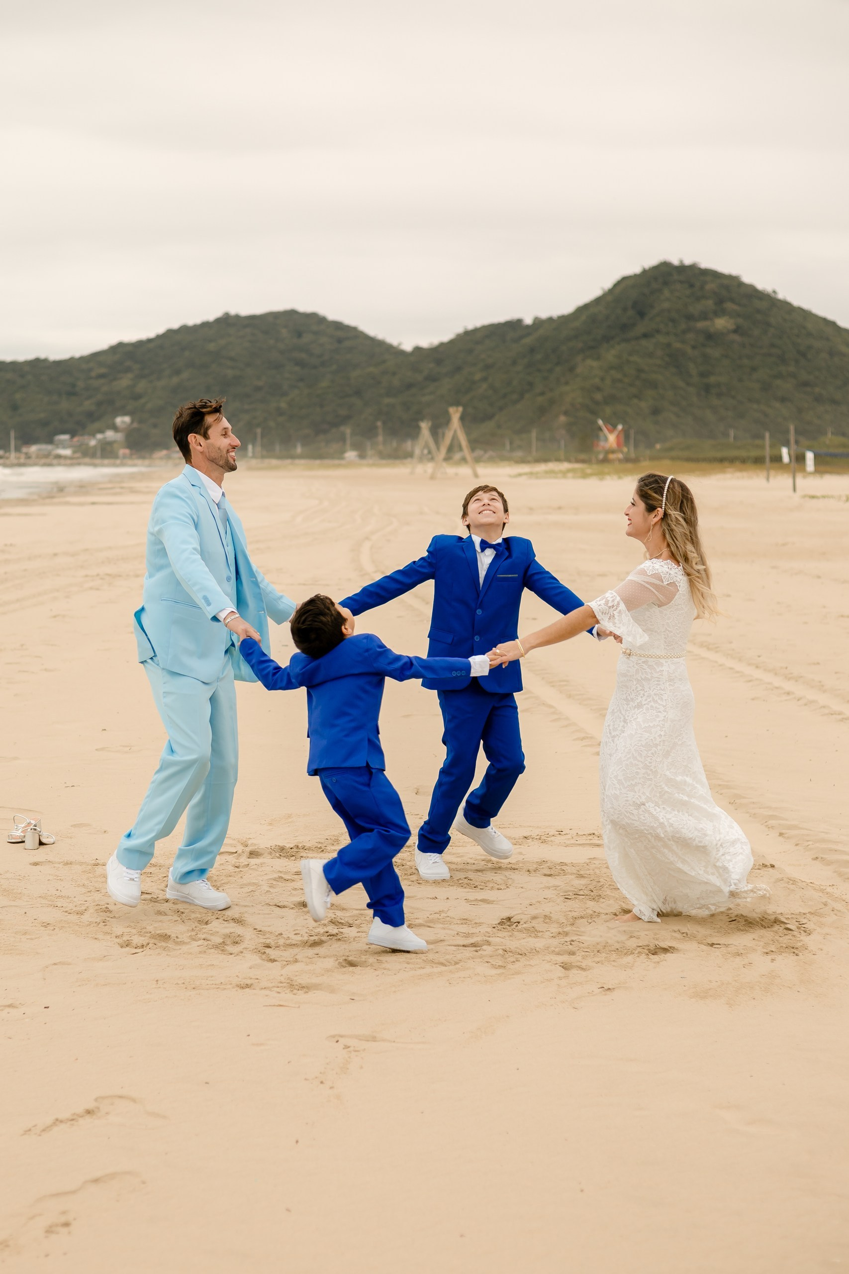 Fotógrafo de casamento em Navegantes; a família de mãos dadas formando uma roda alegre na areia da praia, tendo os noivos e os dois pajens em um momento descontraído do ensaio externo.
