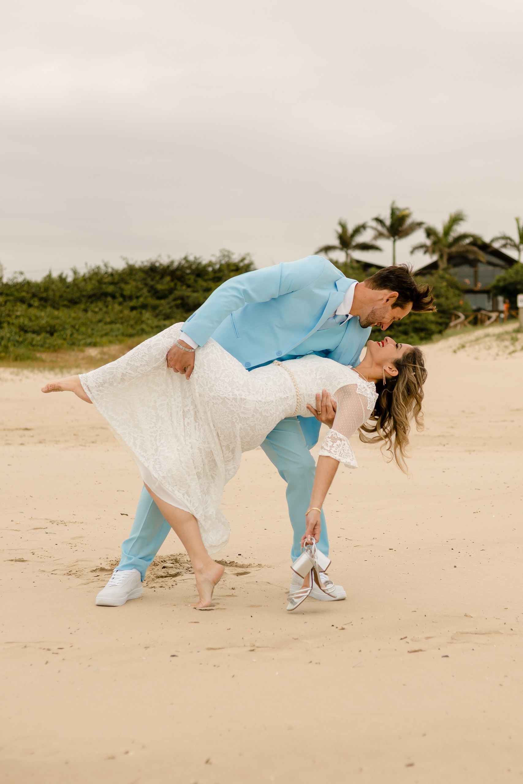 Fotógrafo de casamento em Navegantes; o noivo inclina a noiva para um beijo romântico na areia da praia durante o ensaio externo.