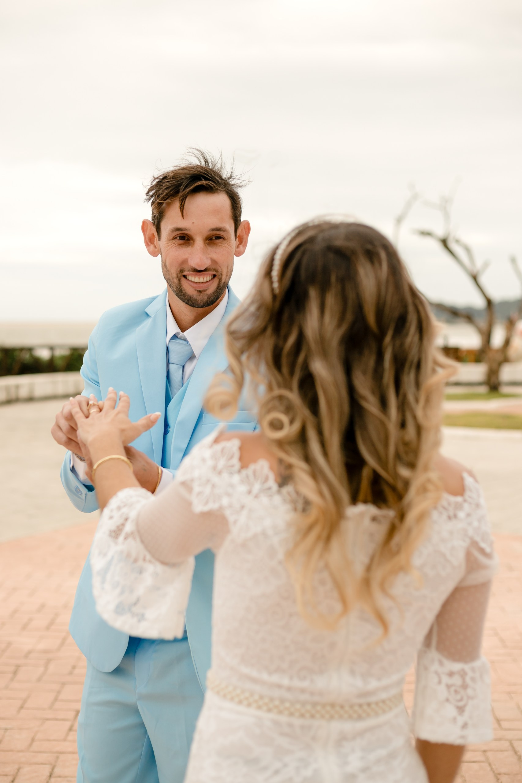 Fotógrafo de casamento em Navegantes; o noivo sorri olhando para a noiva (em primeiro plano de costas) durante ensaio fotográfico externo na orla da praia.