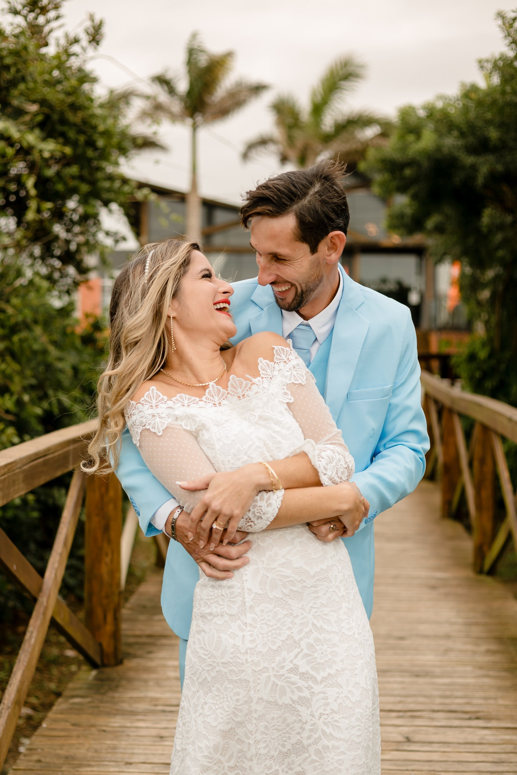 Fotógrafo de casamento em Navegantes; o noivo abraça a noiva por trás, ambos rindo alegremente sobre uma ponte de madeira durante ensaio externo na praia.