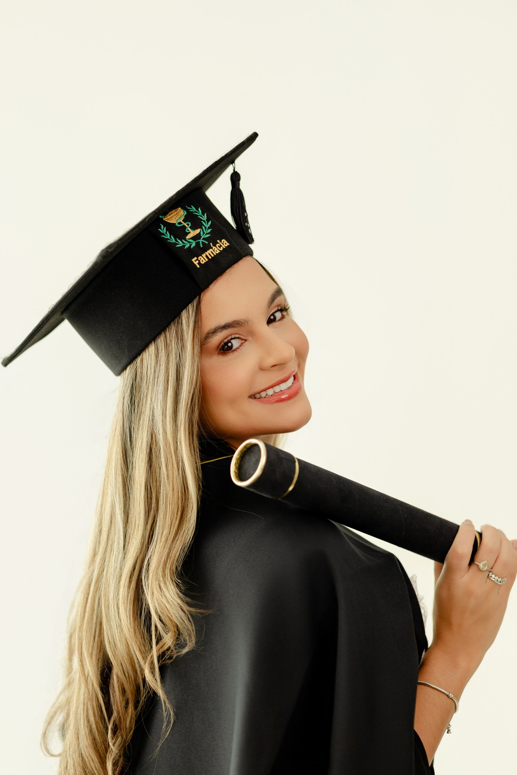 Fotografia de ensaio de formatura em Balneário Camboriú com formanda de farmácia sorrindo por cima do ombro, vestindo capelo com bordado do curso e segurando o canudo.