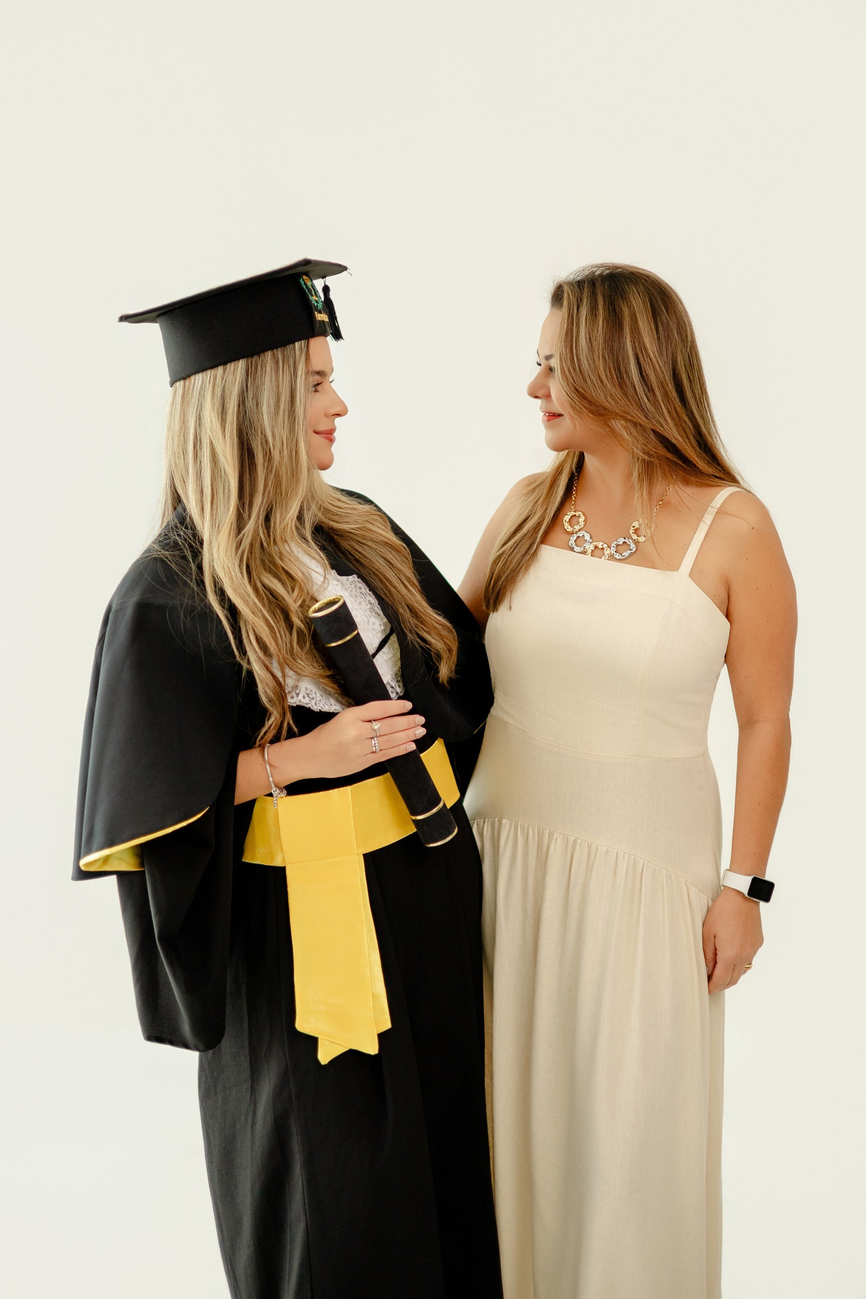 Fotografia de ensaio de formatura em Balneário Camboriú com formanda de farmácia em beca e canudo sorrindo ao lado da mãe com vestido claro.