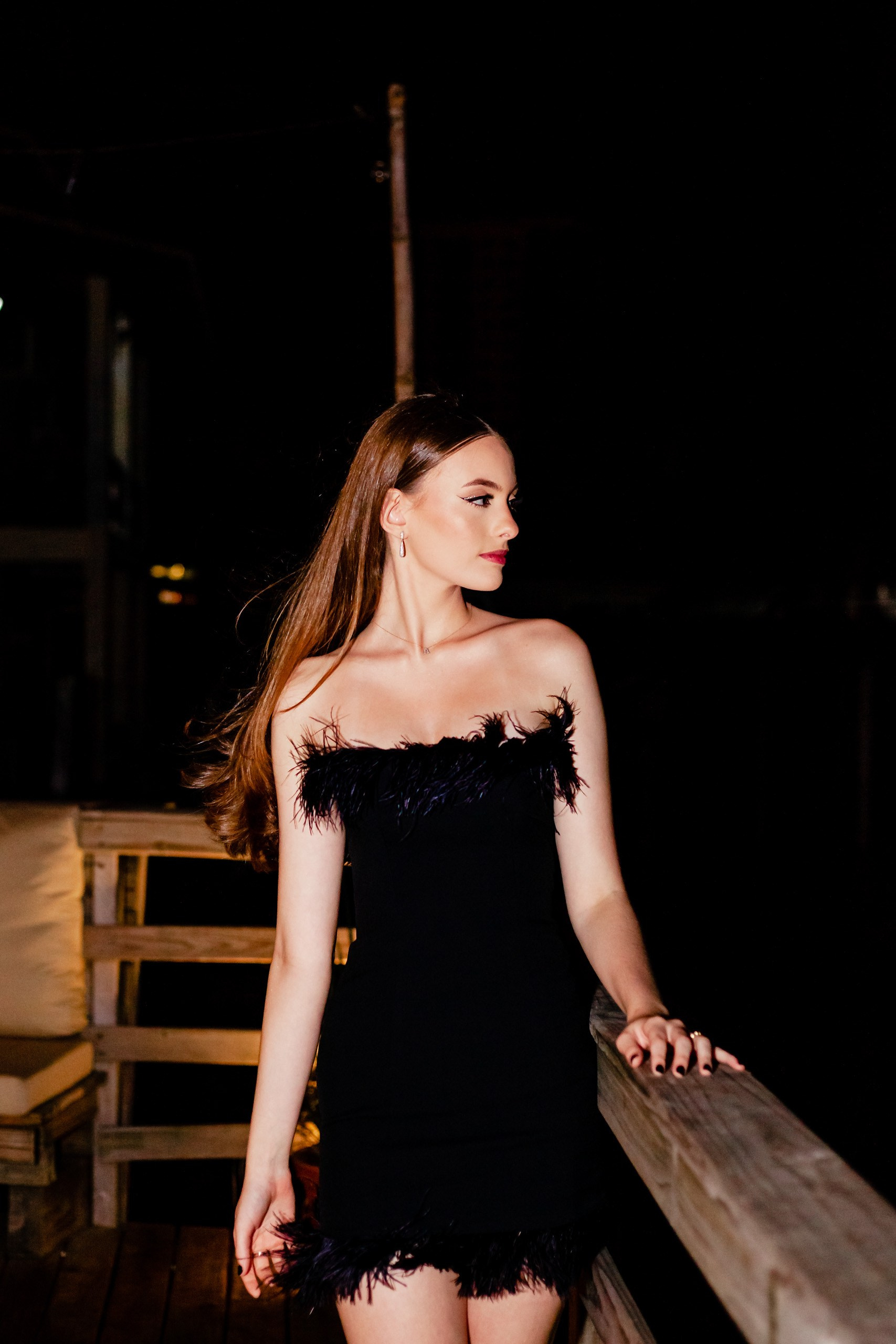 Fotógrafo festa de aniversário em Itajaí; retrato individual elegante da aniversariante vestida de preto, apoiada em uma estrutura de madeira durante a festa noturna.