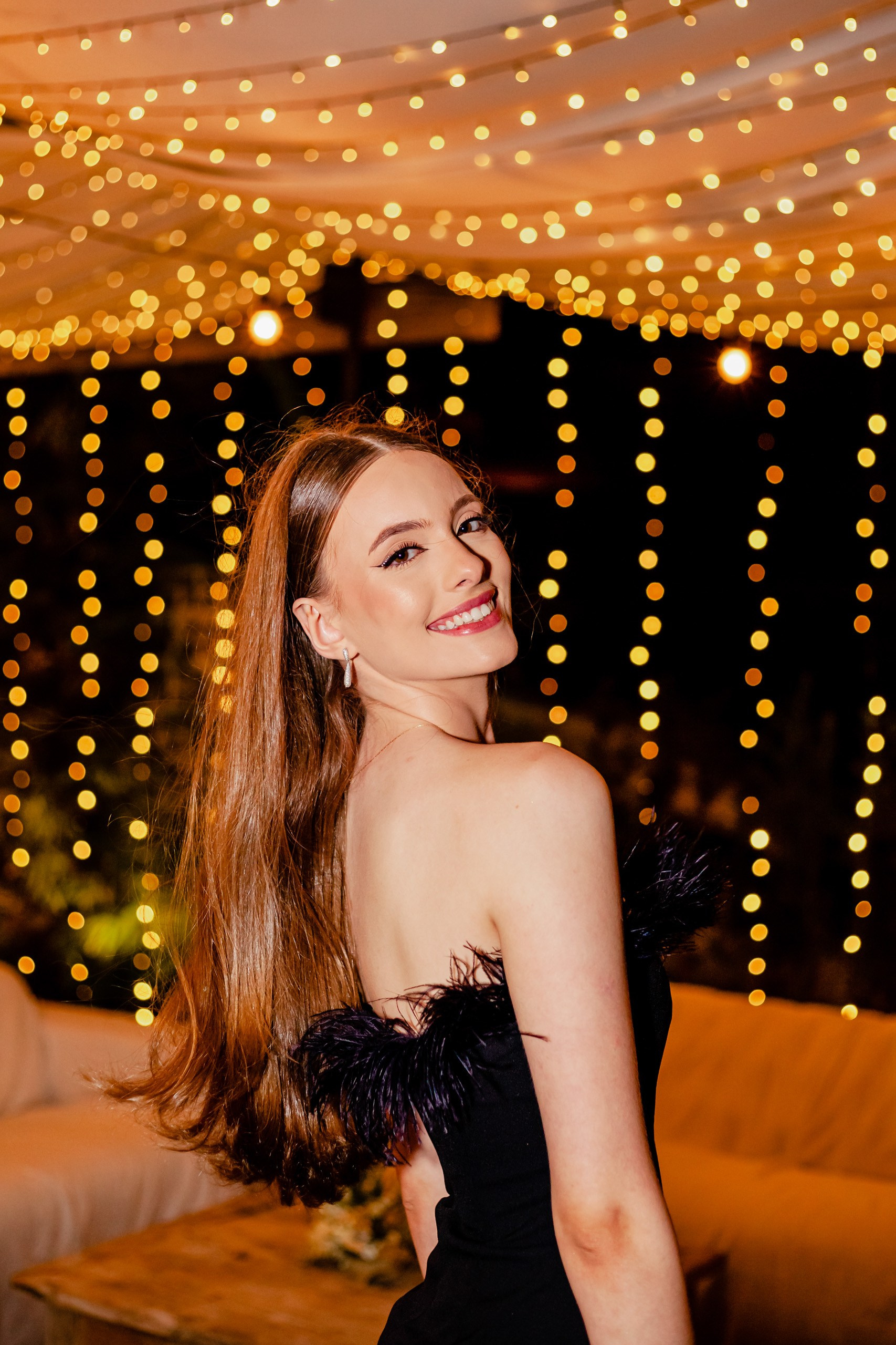 Fotógrafo festa de aniversário em Itajaí; retrato elegante e sorridente da aniversariante olhando por cima do ombro, emoldurada por uma iluminação cênica de luzes pendentes.