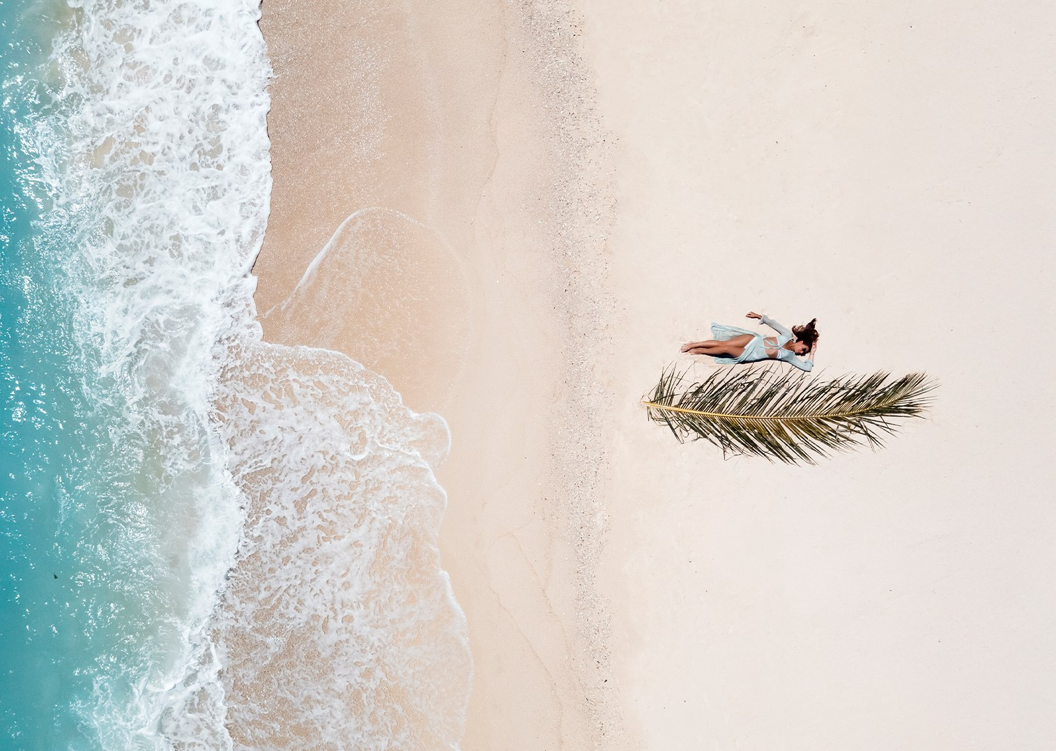 Aerial lifestyle campaign featuring a woman relaxing on tropical sand with a palm lea
