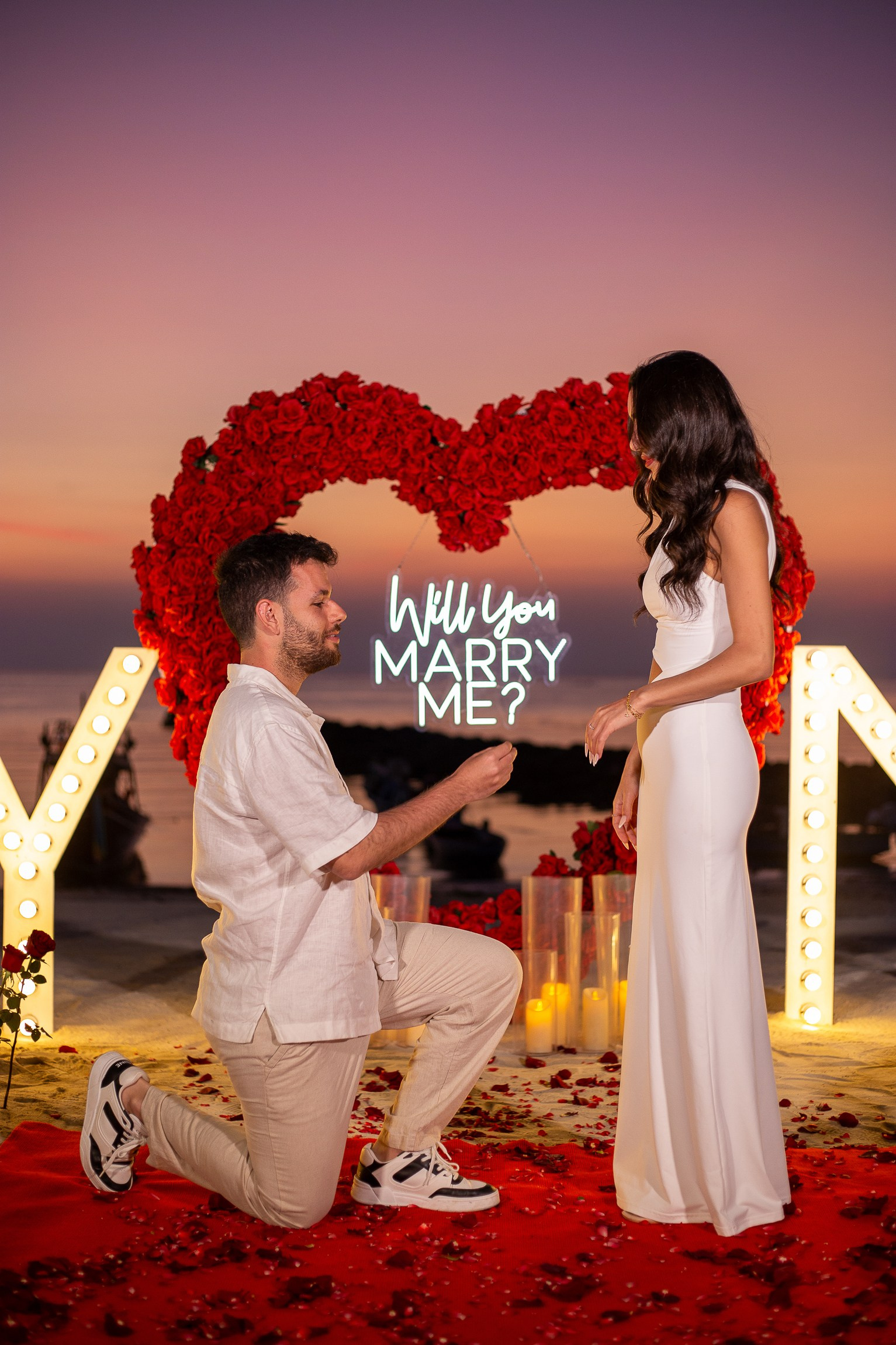 Man proposing on one knee in front of a red floral heart and illuminated “Marry Me” decorations in Pattaya.