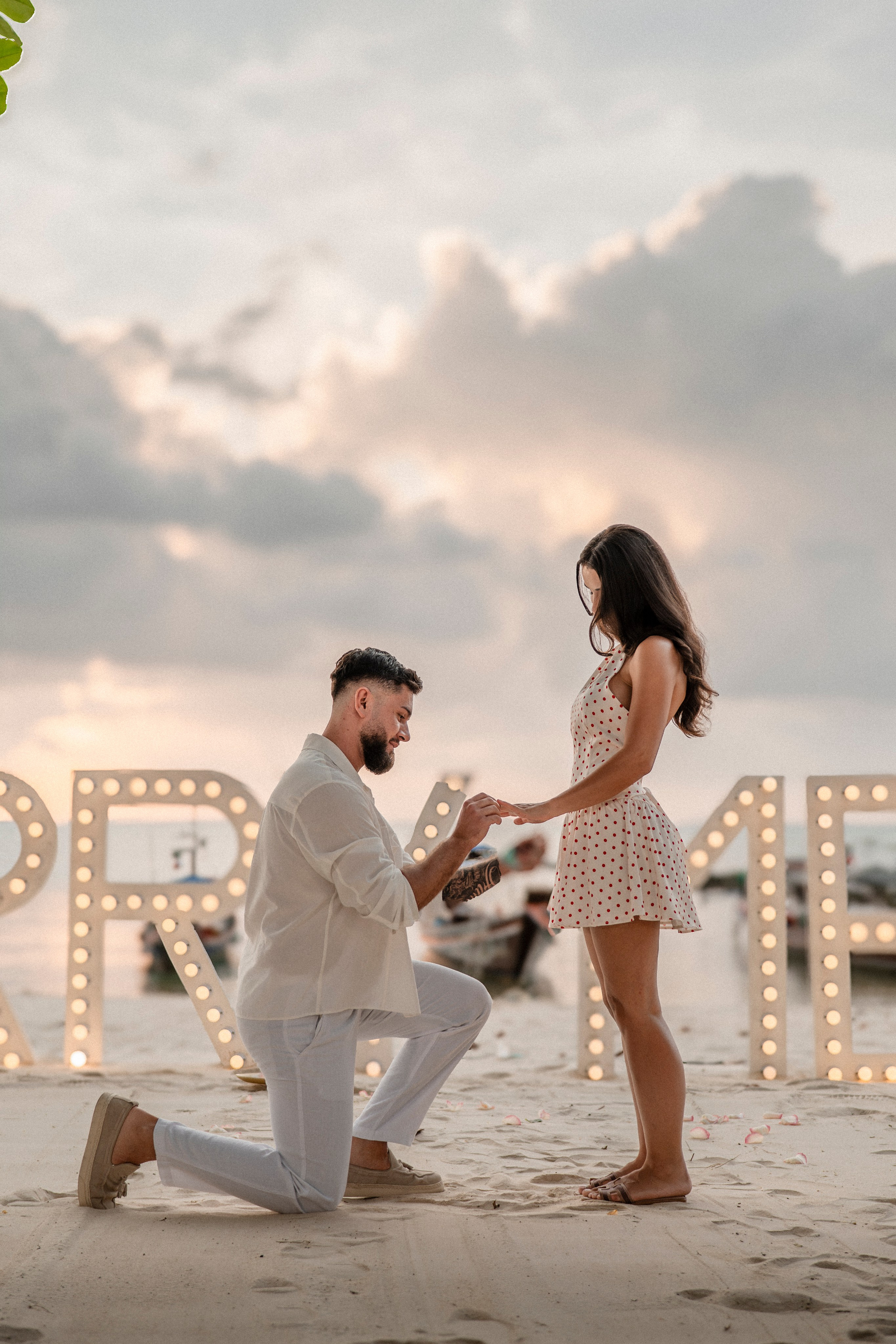 Man proposing on one knee to his partner on a Pattaya beach with illuminated “Marry Me” letters in the background.