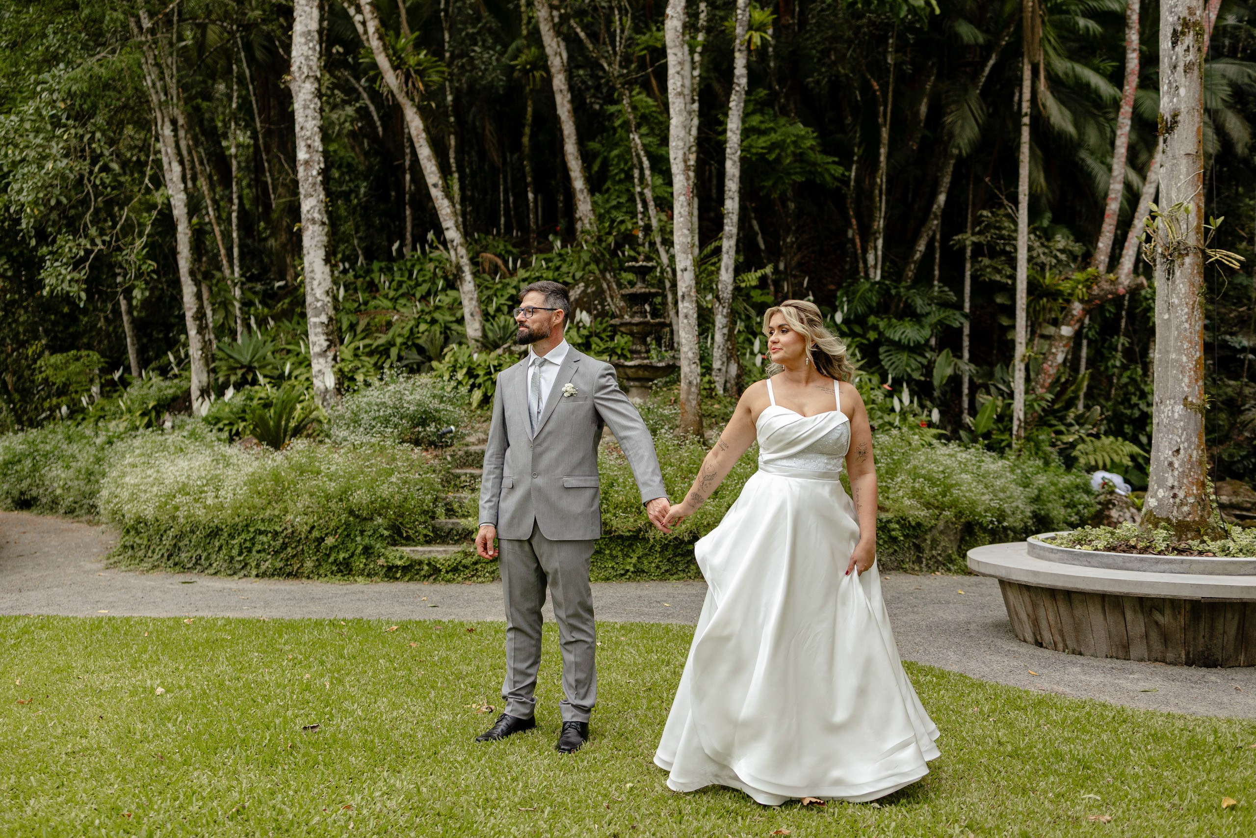 Fotógrafo de casamento em Santa Catarina; noivos de mãos dadas de pé na grama em jardim arborizado e natural durante ensaio ao ar livre.