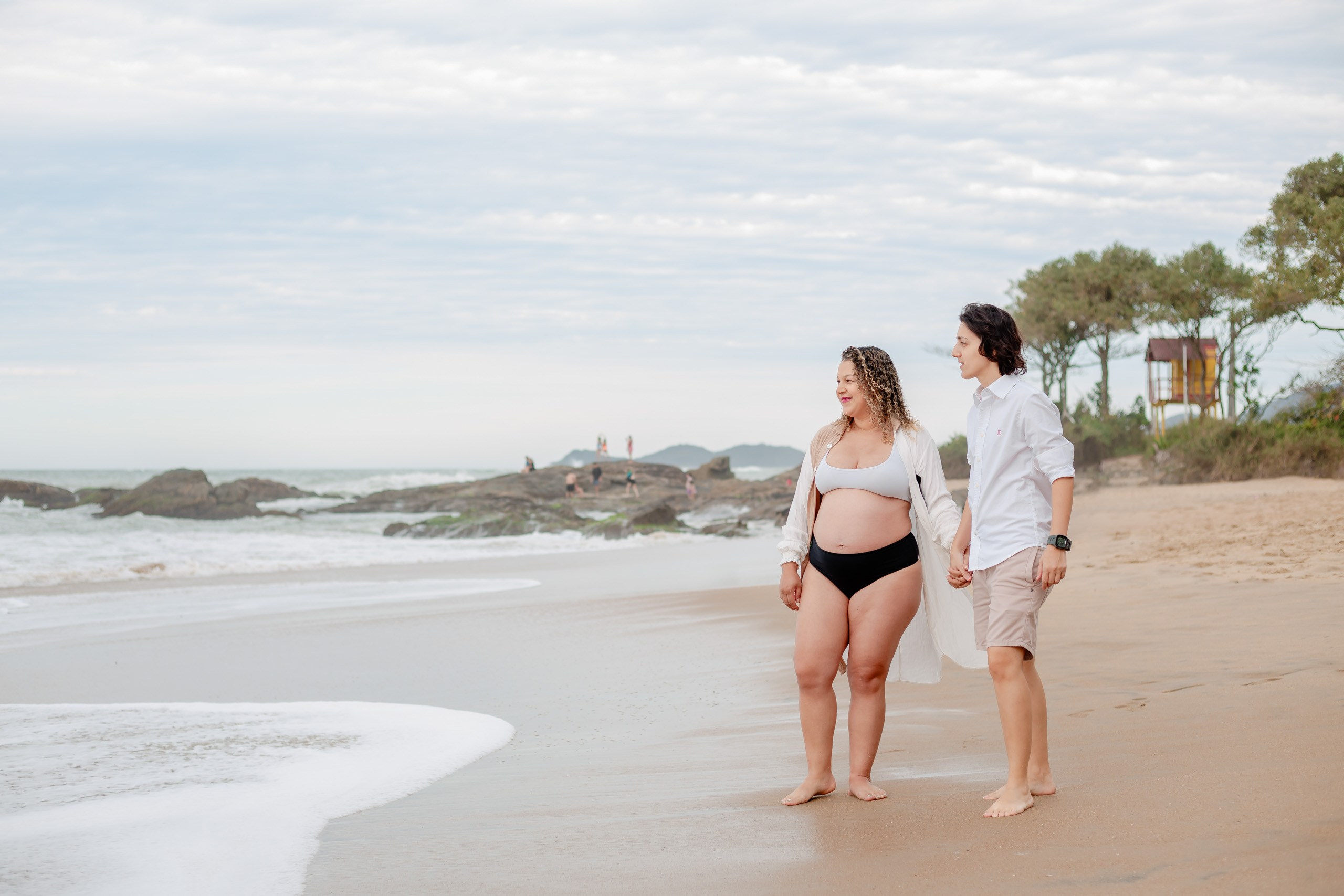 Fotógrafo ensaio gestante em Penha; plano geral do casal caminhando e contemplando a paisagem na praia, em um ensaio lifestyle leve e natural à beira-mar.