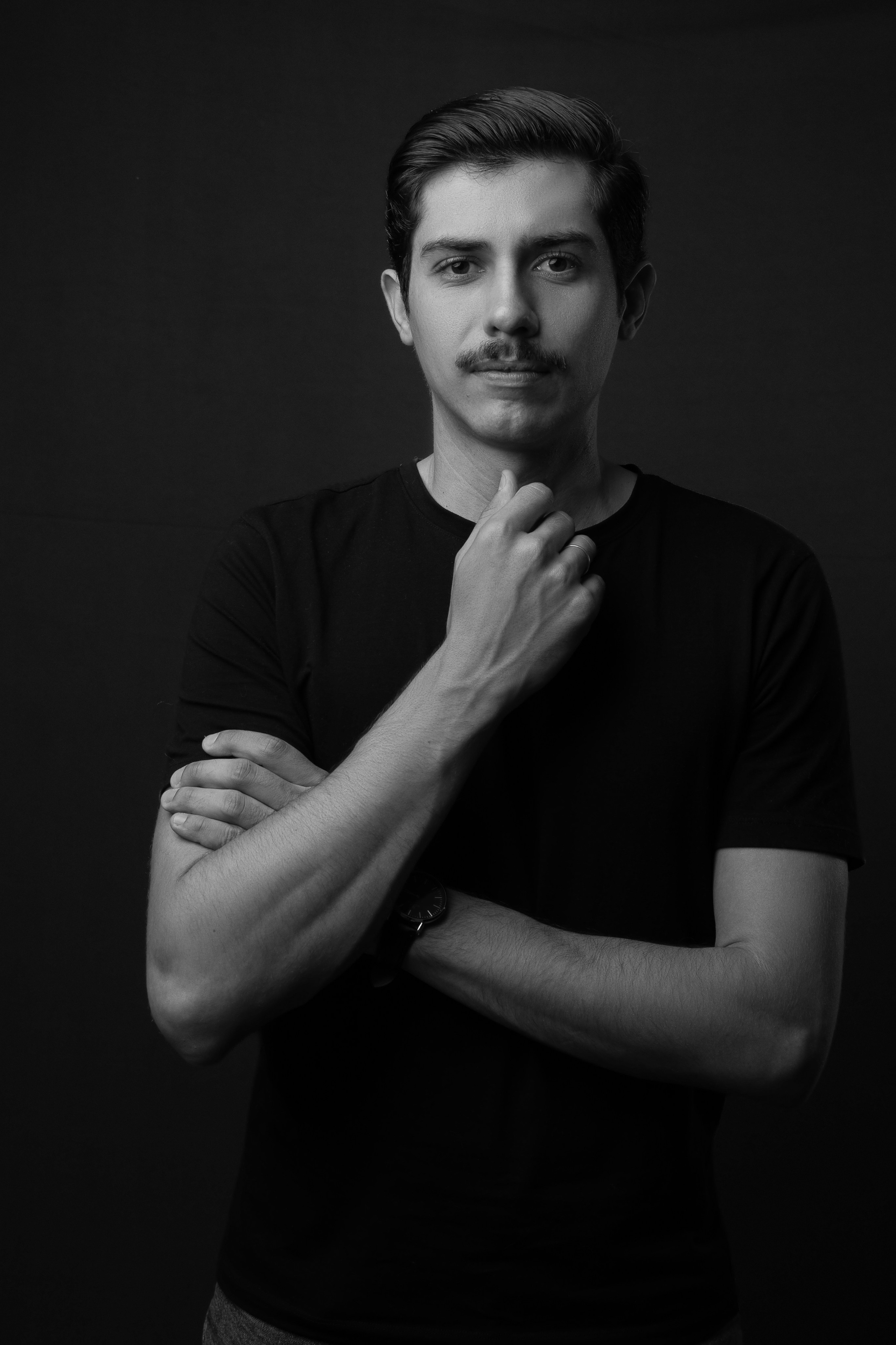 Retrato corporativo em estúdio de fisioterapeuta vestindo camiseta preta, com braços cruzados sobre fundo escuro.
