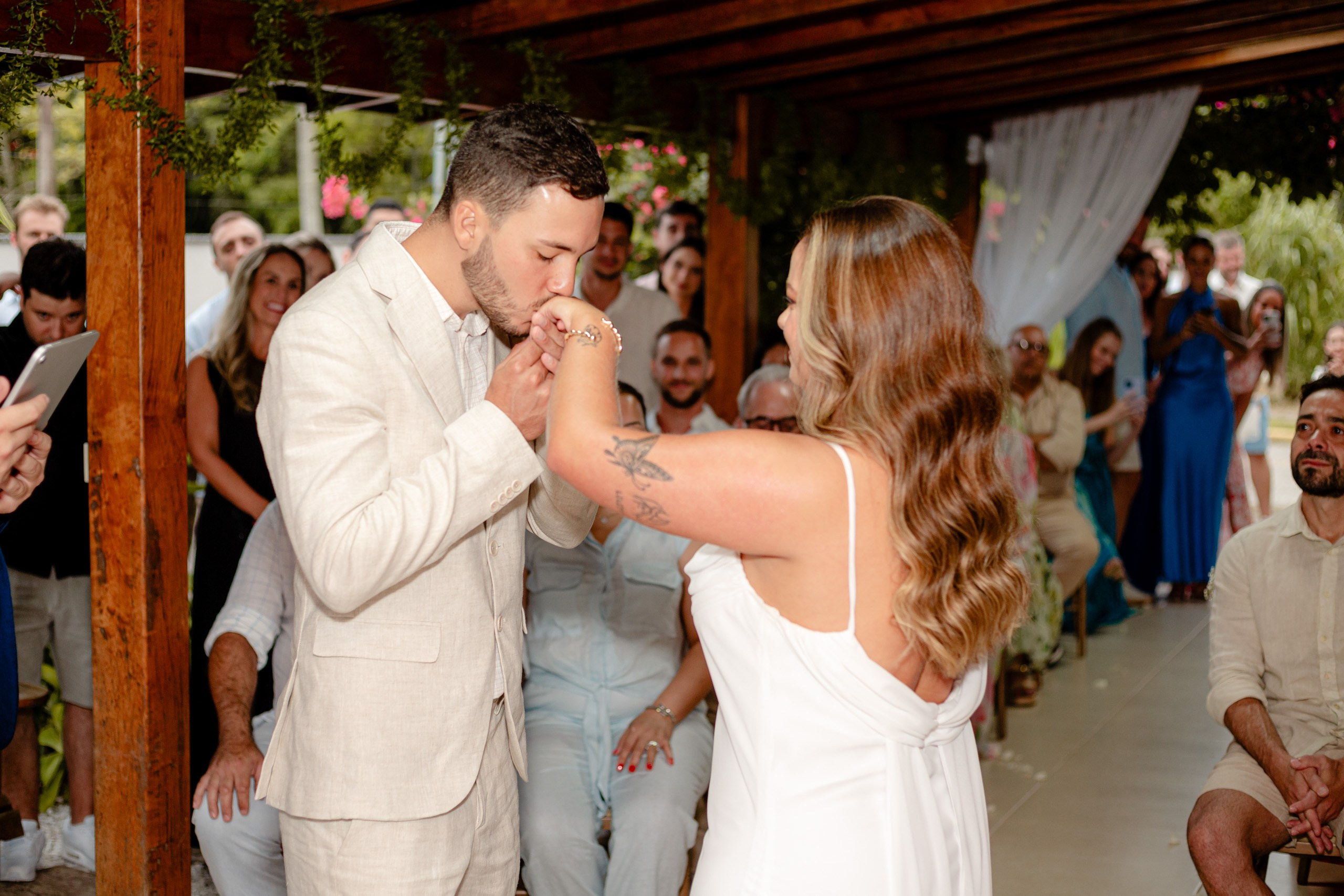 Fotógrafo de casamento em Bombinhas; o noivo beija delicadamente a mão da noiva durante a cerimônia sob a estrutura de madeira.