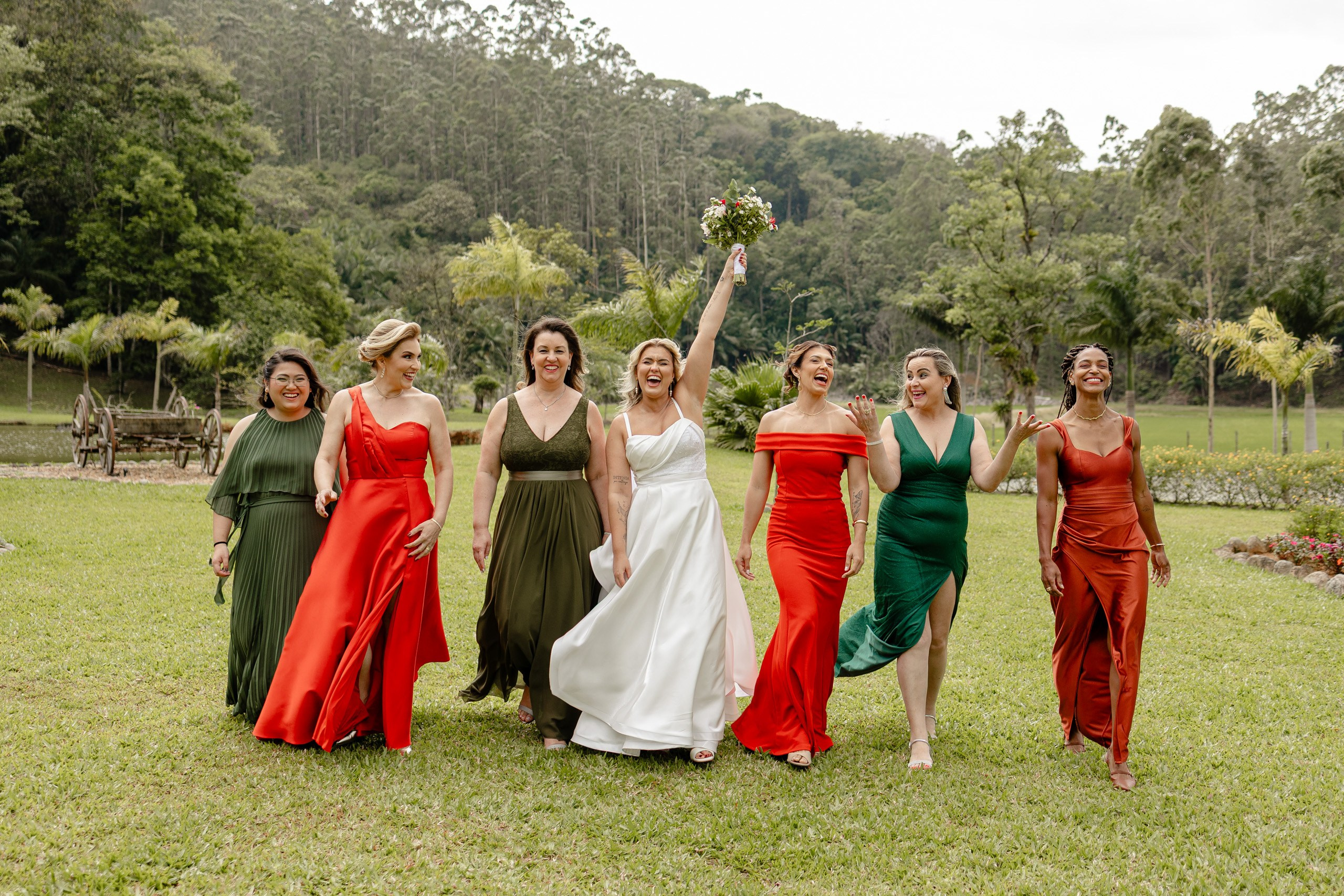 Fotógrafo de casamento em Balneário Camboriú; retrato dinâmico e divertido da noiva com seu vestido branco ao centro, caminhando em um gramado ao ar livre junto com suas madrinhas vestidas em elegantes tons de verde e vermelho.
