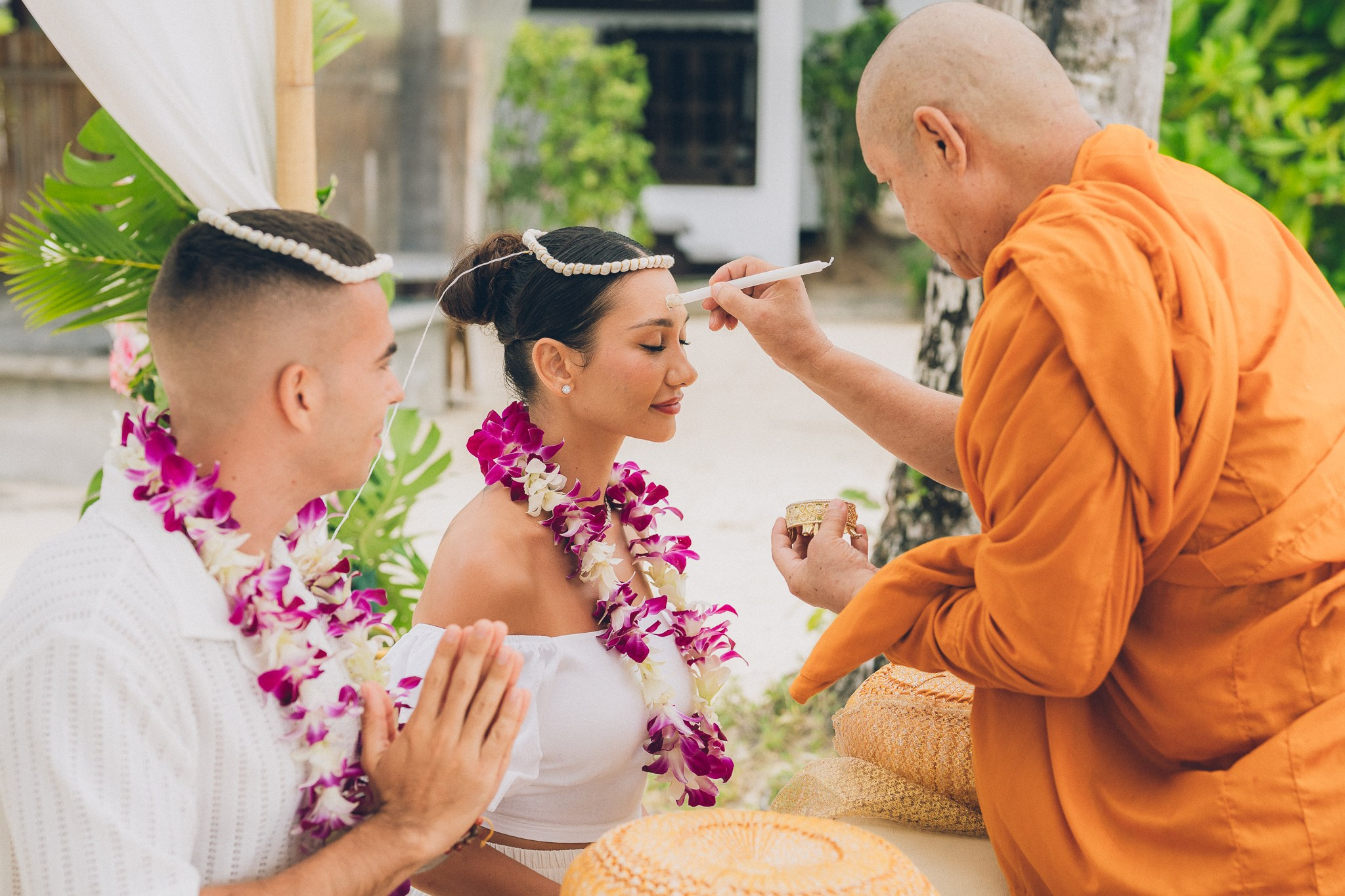 Traditional Thai Buddhist wedding ceremony in Koh Samui, Thailand, with a Buddhist monk blessing the bride and groom wearing white outfits and purple orchid garlands.