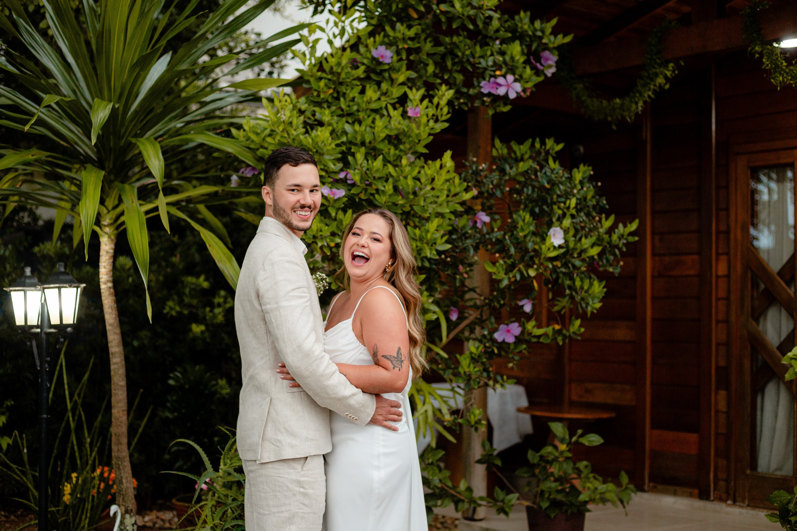 Fotógrafo de casamento em Bombinhas; o noivo sorri enquanto a noiva gargalha de felicidade abraçada a ele durante o ensaio no jardim.