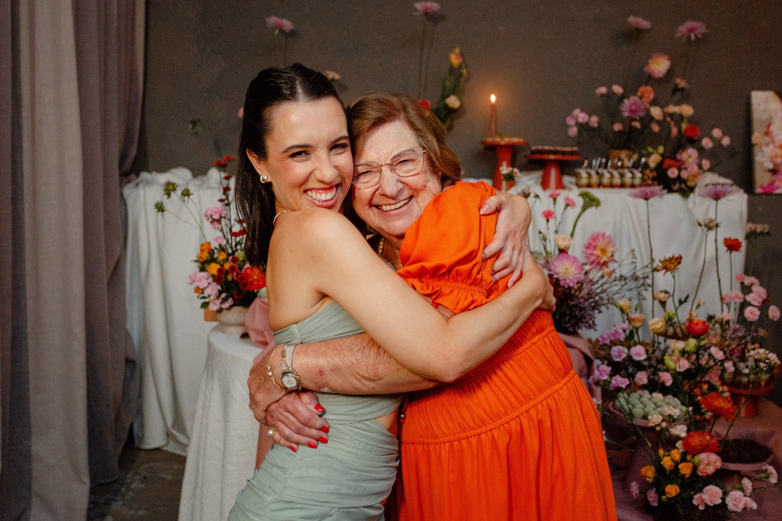 Fotógrafo de festa de formatura em Balneário Camboriú; formanda sorridente abraçando carinhosamente a avó com vestido laranja diante da mesa decorada.