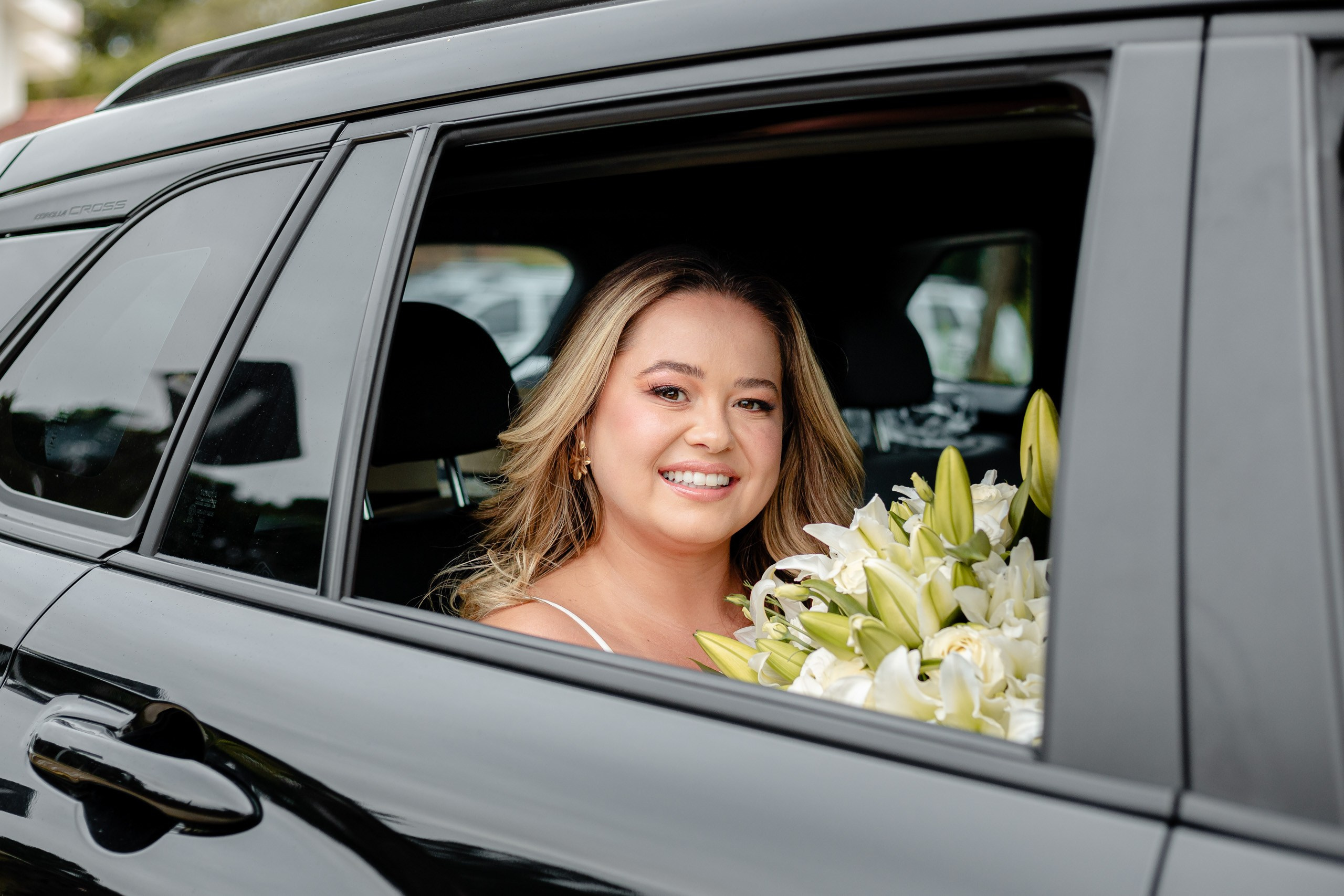 Fotógrafo de casamento em Balneário Camboriú; retrato sorridente da noiva a olhar pela janela de um carro escuro, segurando um elegante buquê de lírios brancos antes da cerimônia.